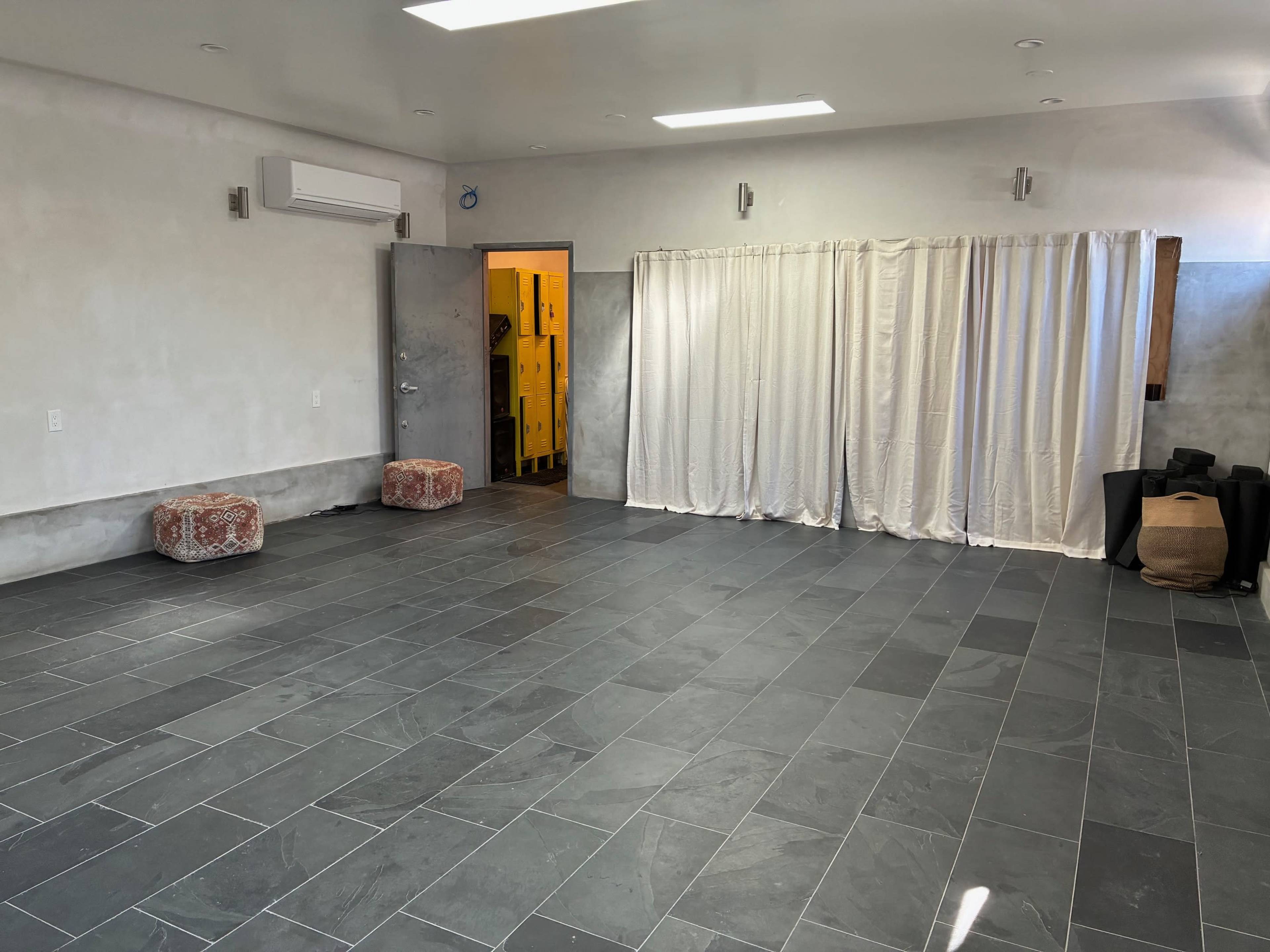 The image shows a spacious, minimalist interior with gray tile flooring, a partition made of white curtains, and two small patterned ottomans.