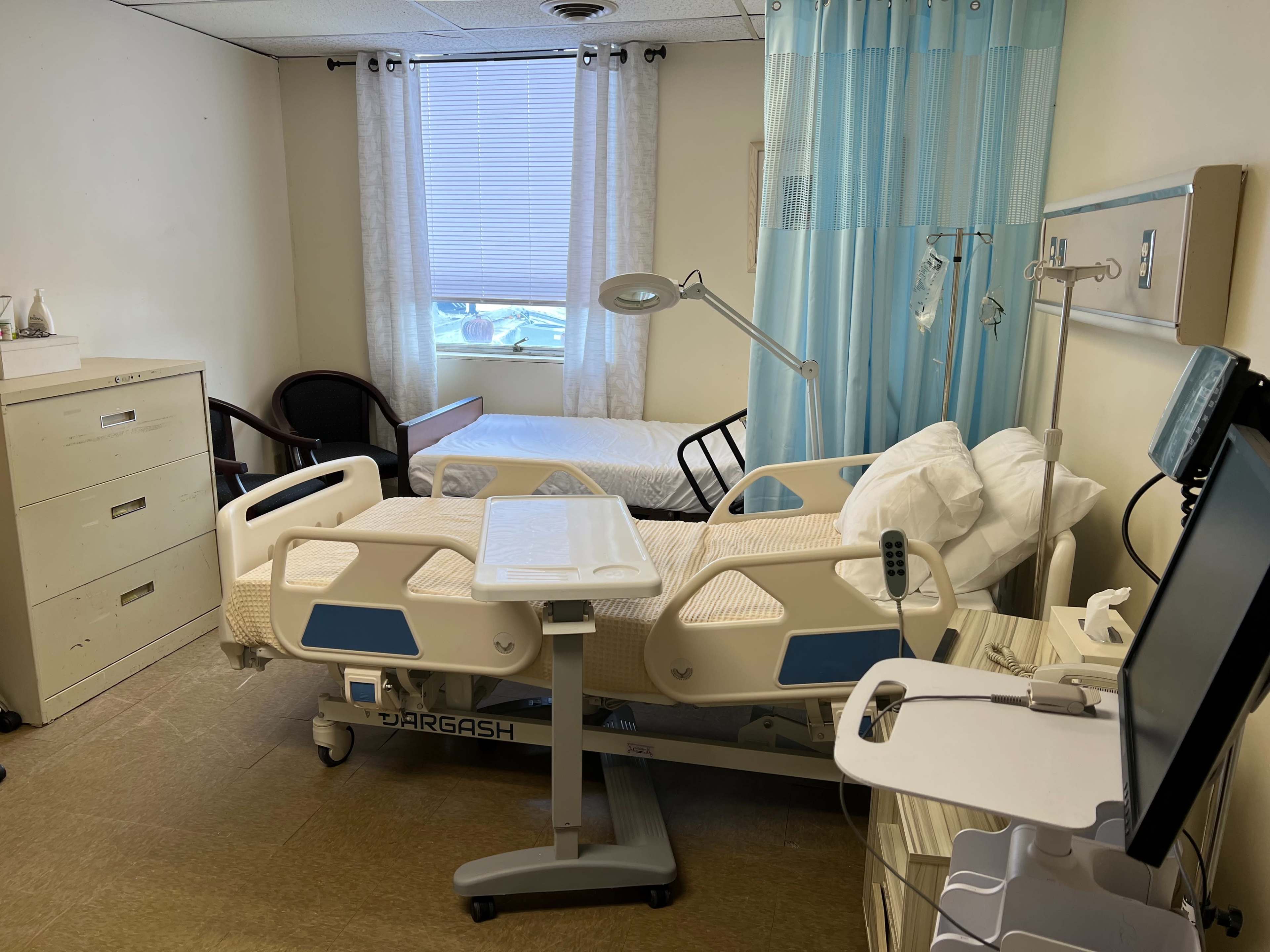 The image shows a hospital room with a bed, a medical supply cart, and a window allowing natural light.