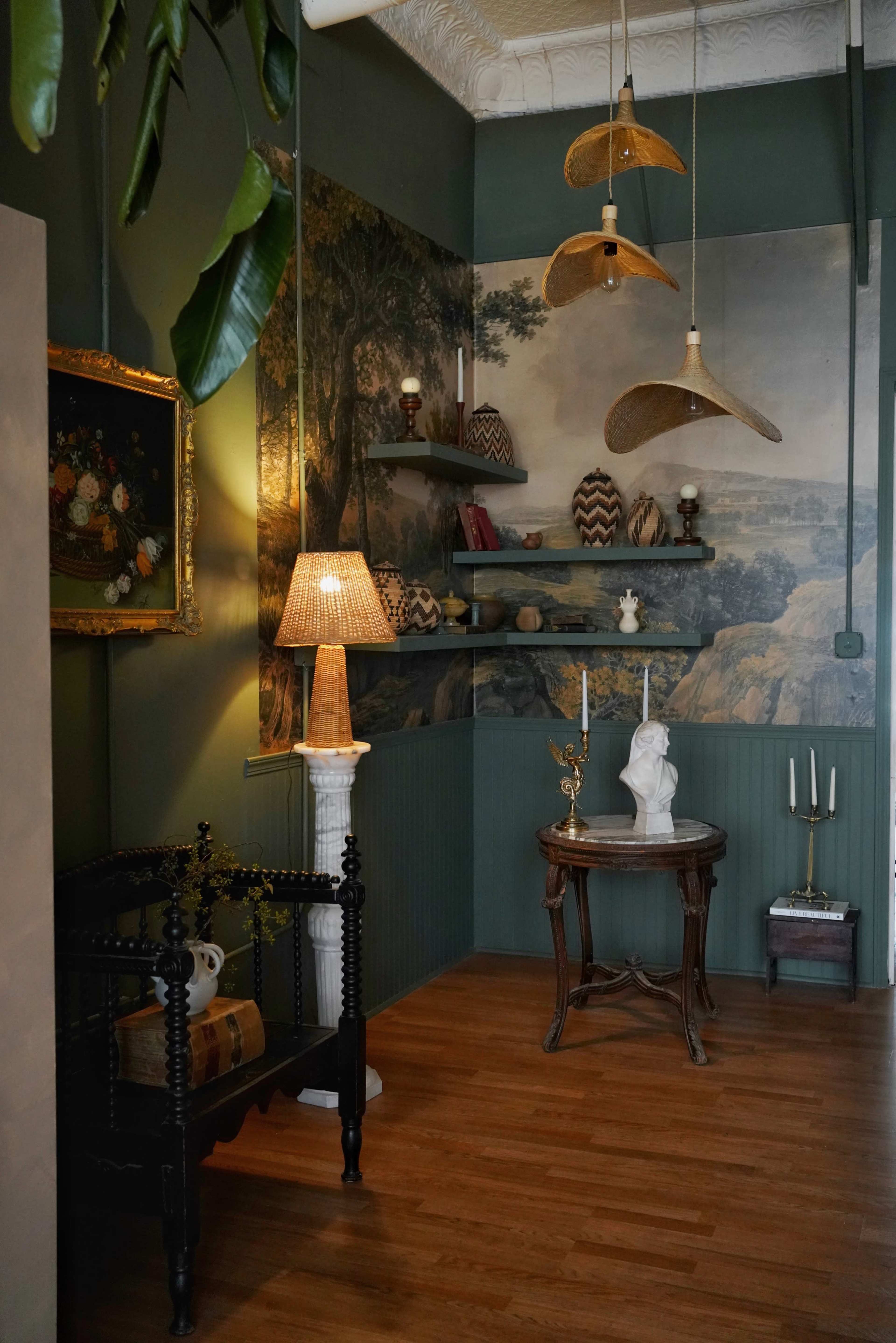 A cozy interior corner featuring a wooden floor, a vintage lamp on a column, various shelves with decorative items, and a statue on a small table under hanging light fixtures.