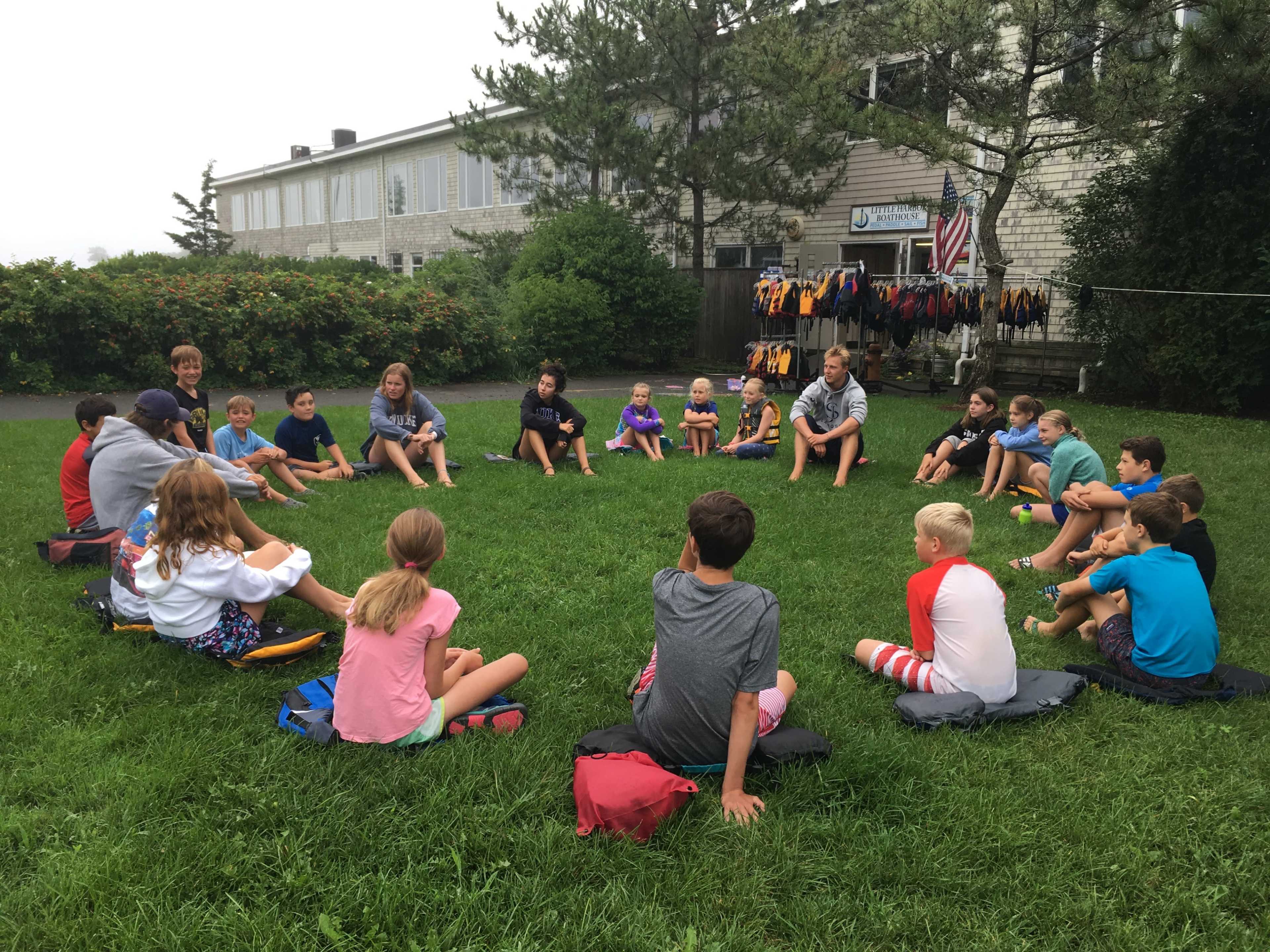 A group of children and adults sits in a circle on the grass in front of a building, engaged in a discussion while life jackets hang on a nearby line.