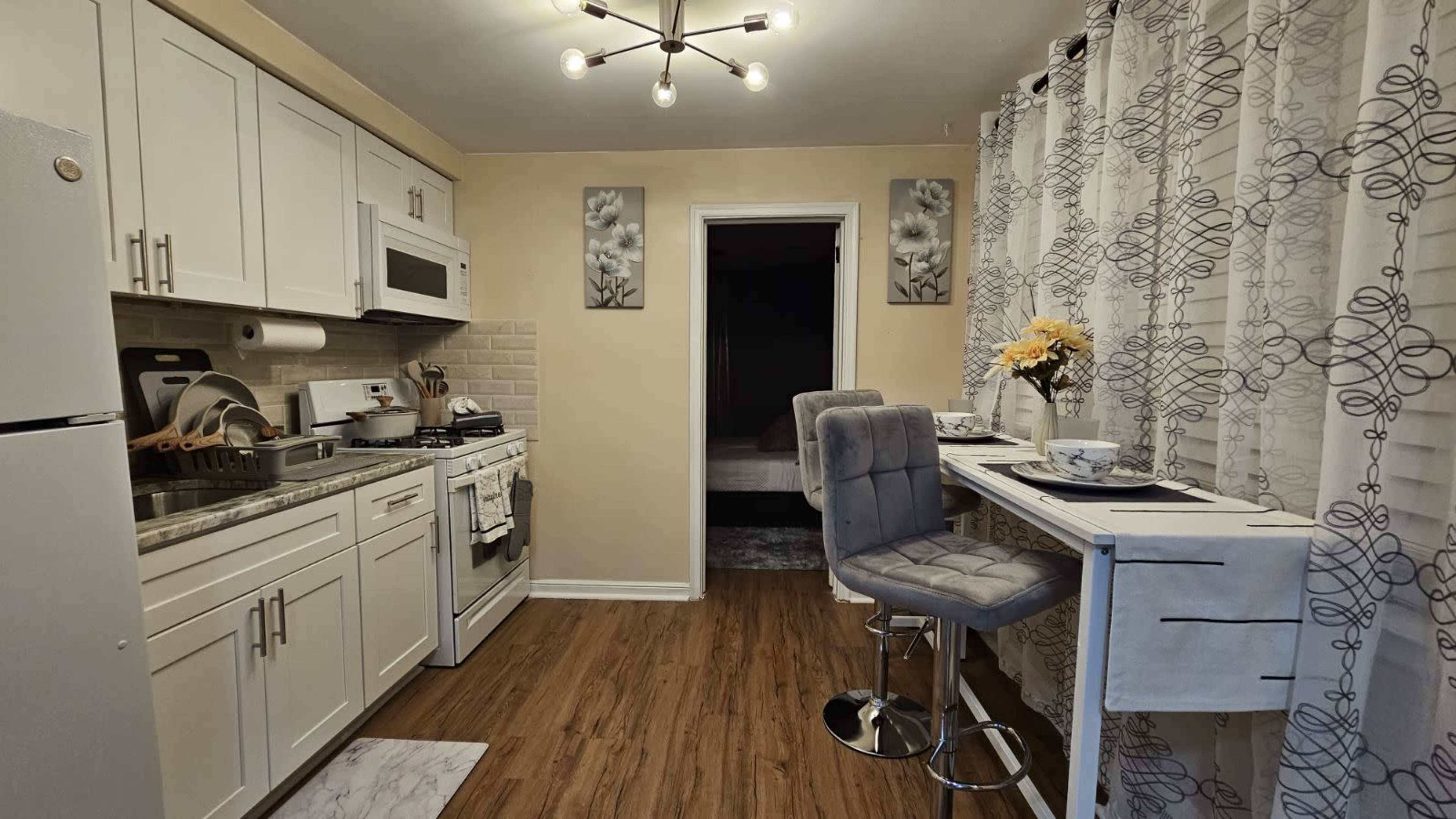 A modern kitchen with white cabinetry, a grey countertop, a small dining area for two, and a doorway leading to a darker room.