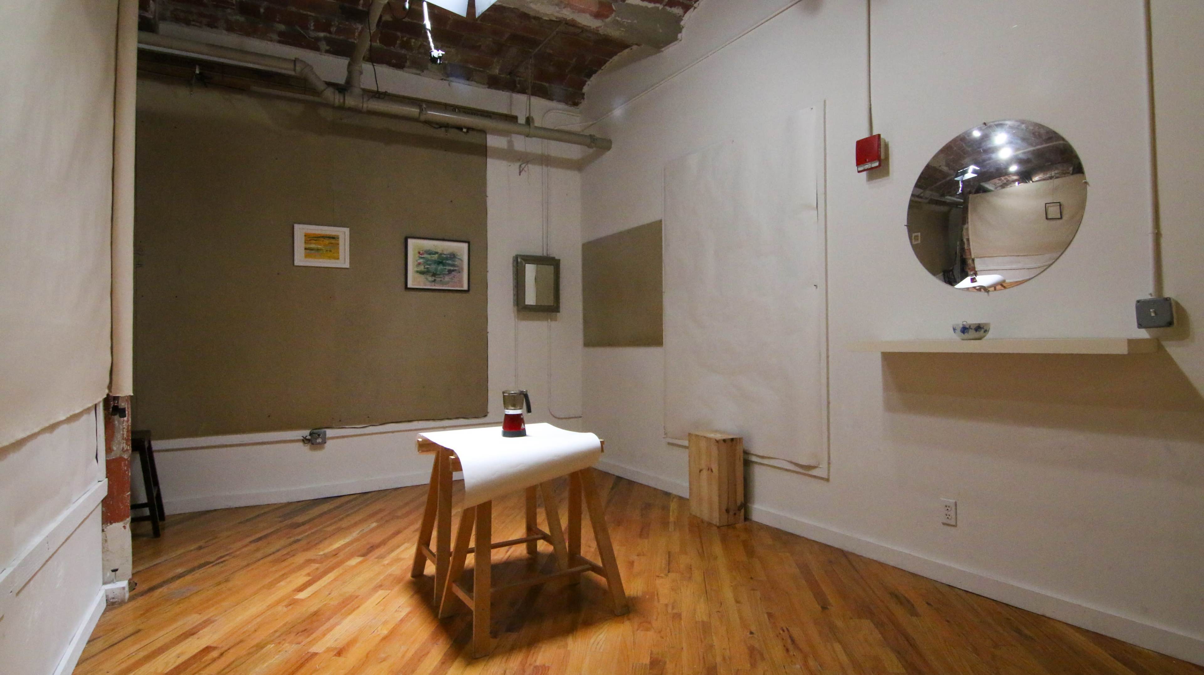 The image shows a small, minimalist room with wooden flooring, featuring a table in the center, a mirror on the wall, and a couple of framed artworks.