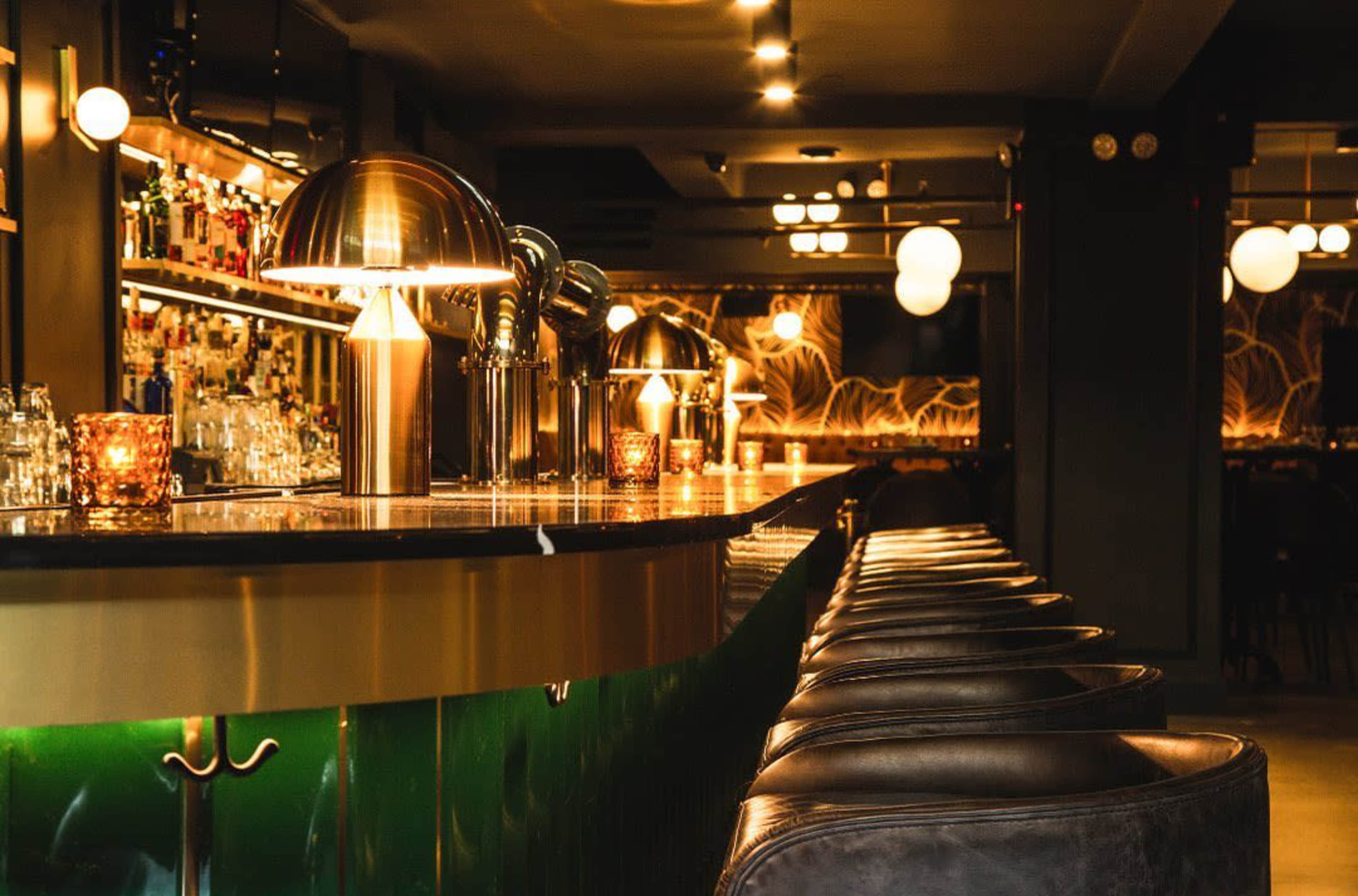 The image shows a sleek bar area with a polished countertop, stylish lighting, and bar stools arranged along the length of the bar.