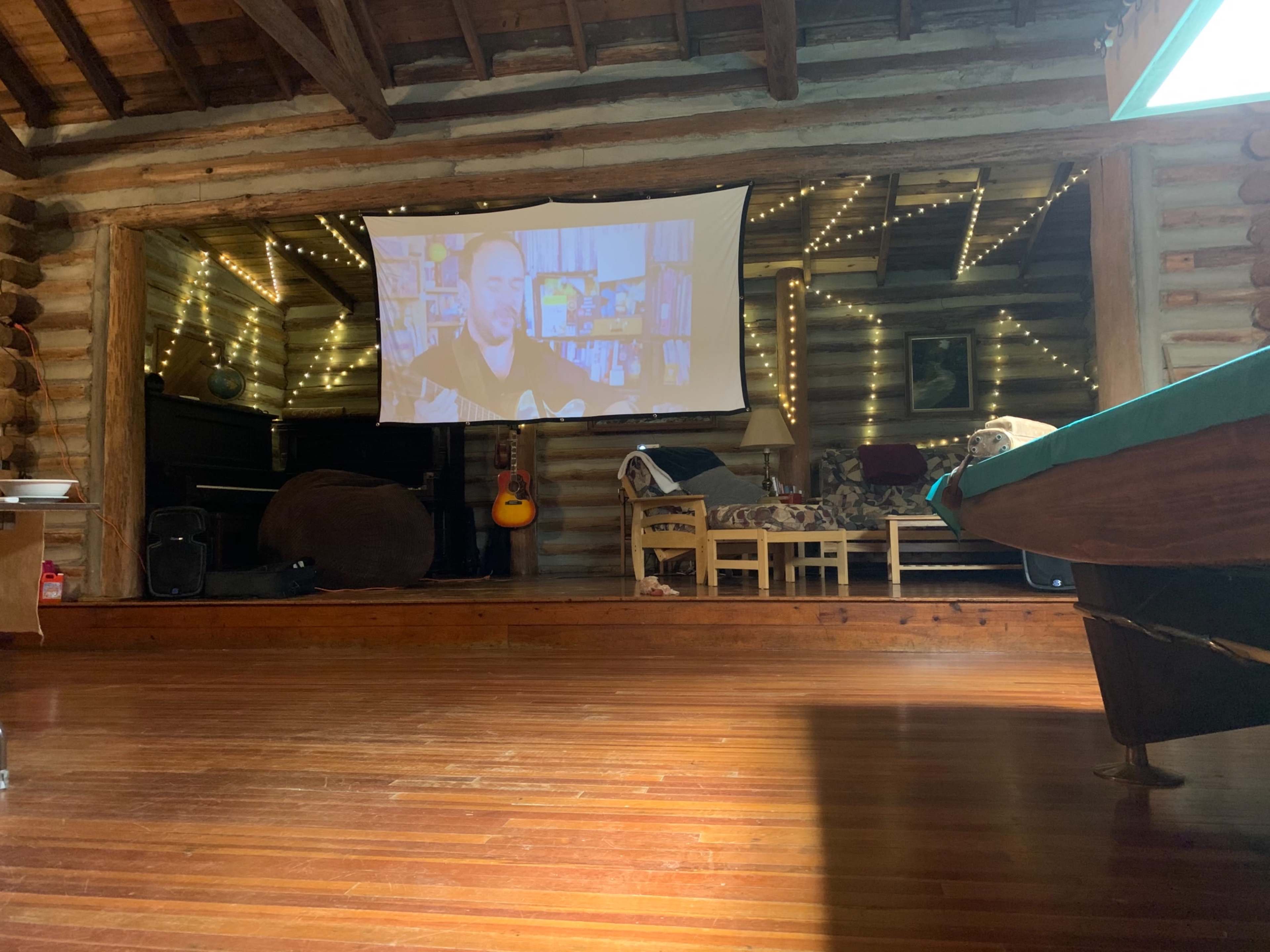 A large screen displays a video in a log cabin setting with wooden walls and string lights, featuring a relaxed seating area.