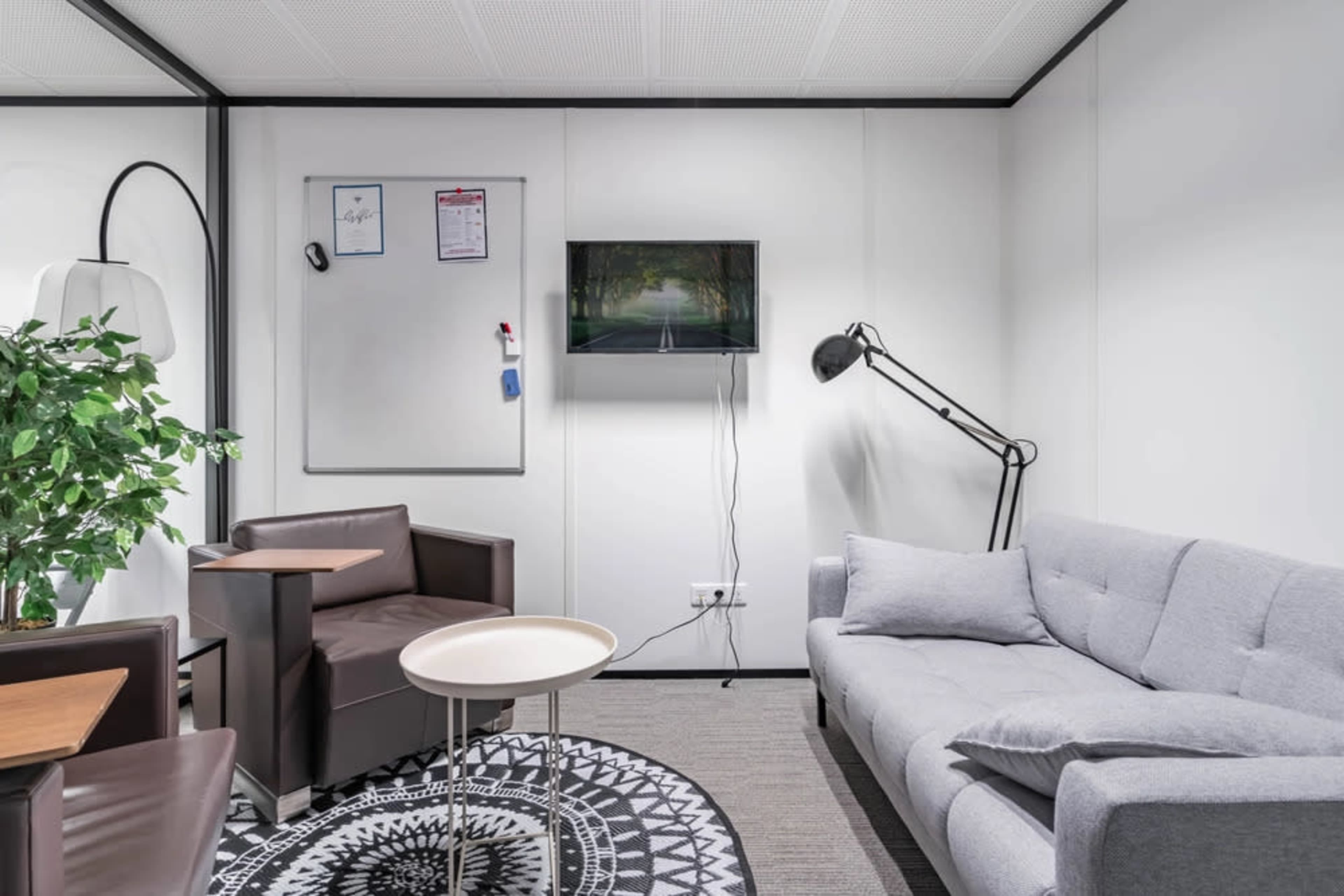 The image shows a modern meeting room featuring a gray couch, two armchairs, a small round table, a floor lamp, a whiteboard, and a television mounted on the wall.