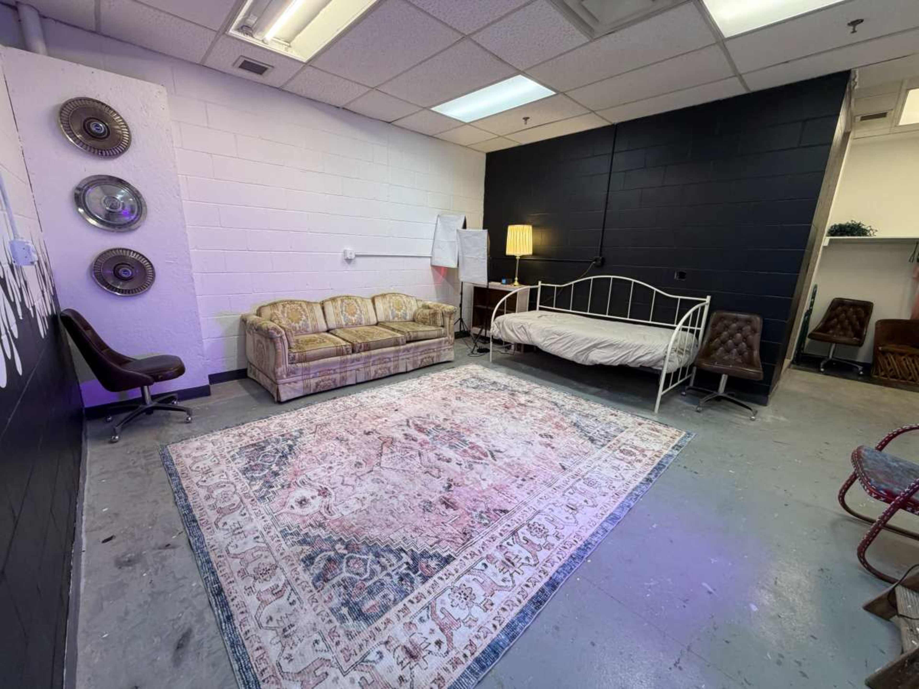 The image shows a room with a beige couch, a white bed, a patterned rug, and a standing lamp against a black wall.