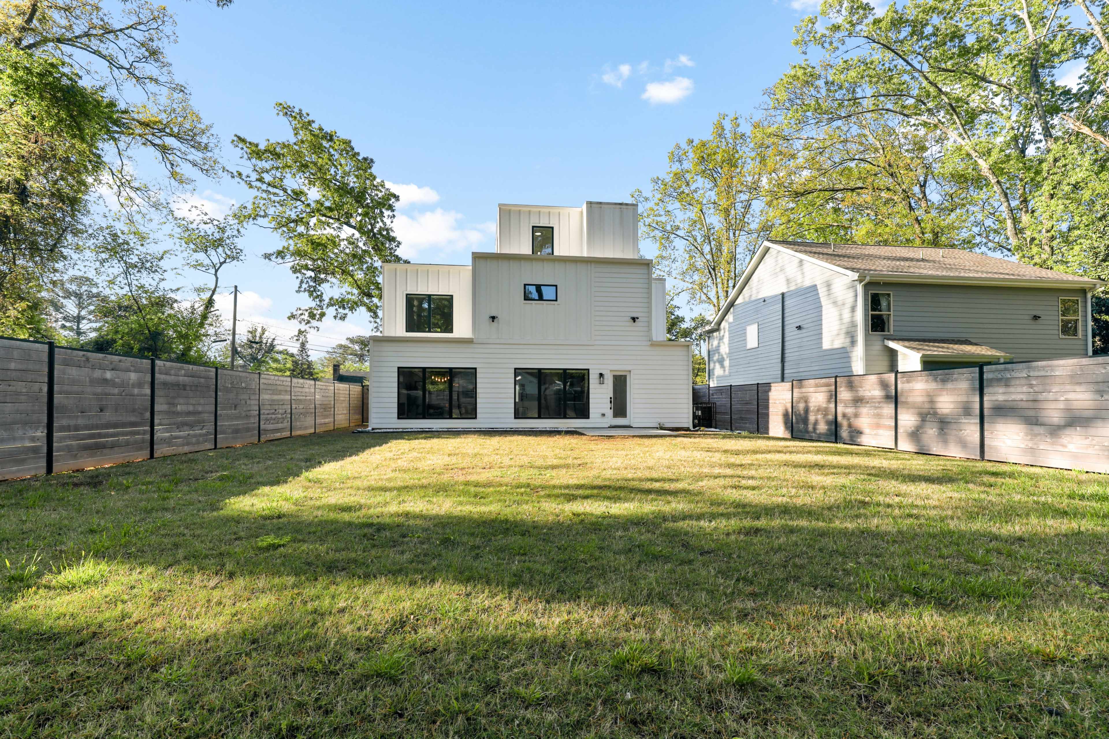 A modern two-story house with large windows is surrounded by a fenced backyard and adjacent homes, set in a grassy area under a clear sky.