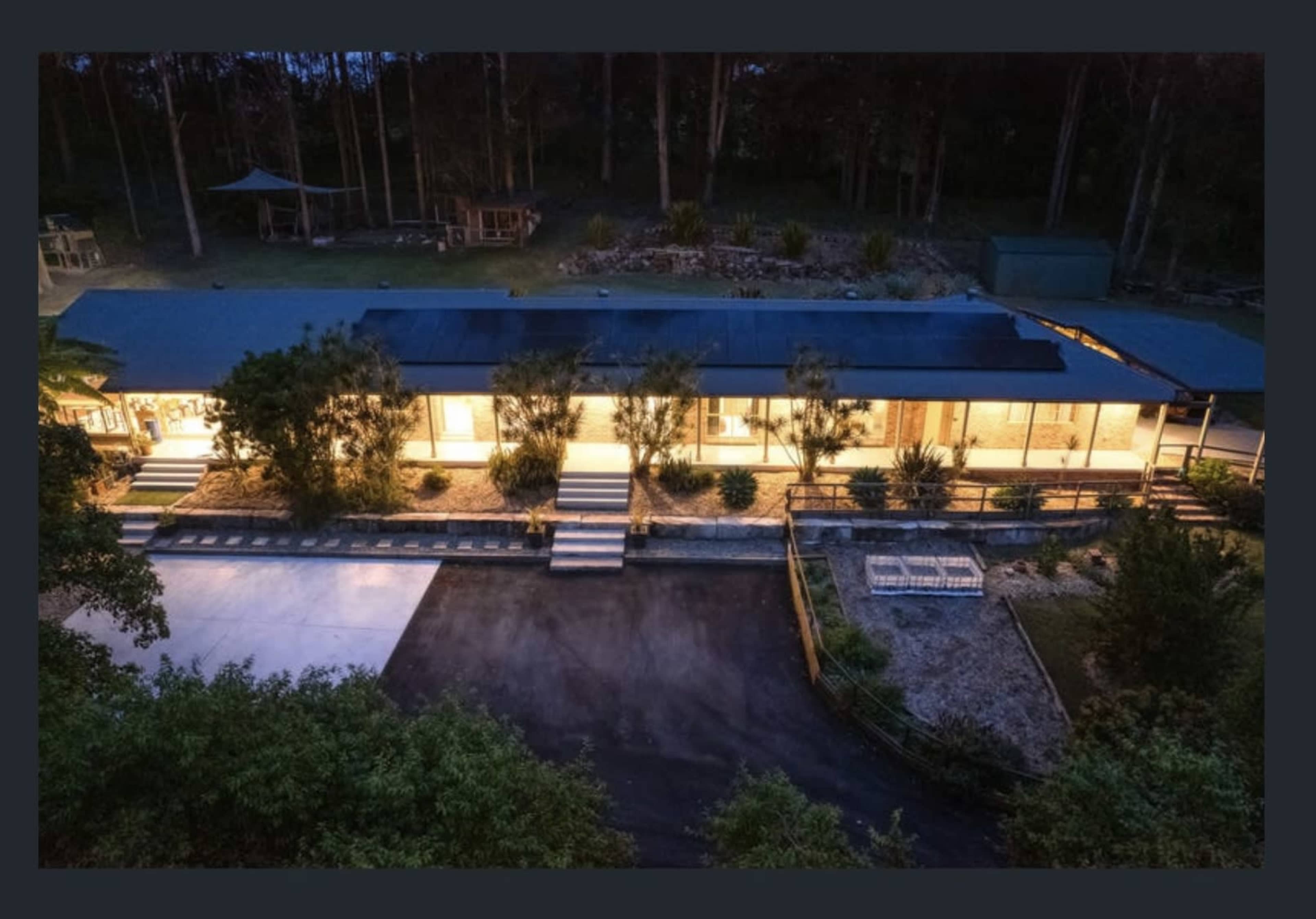 A modern building with illuminated windows sits amidst trees in a dark setting, featuring a driveway and landscaped area in the foreground.