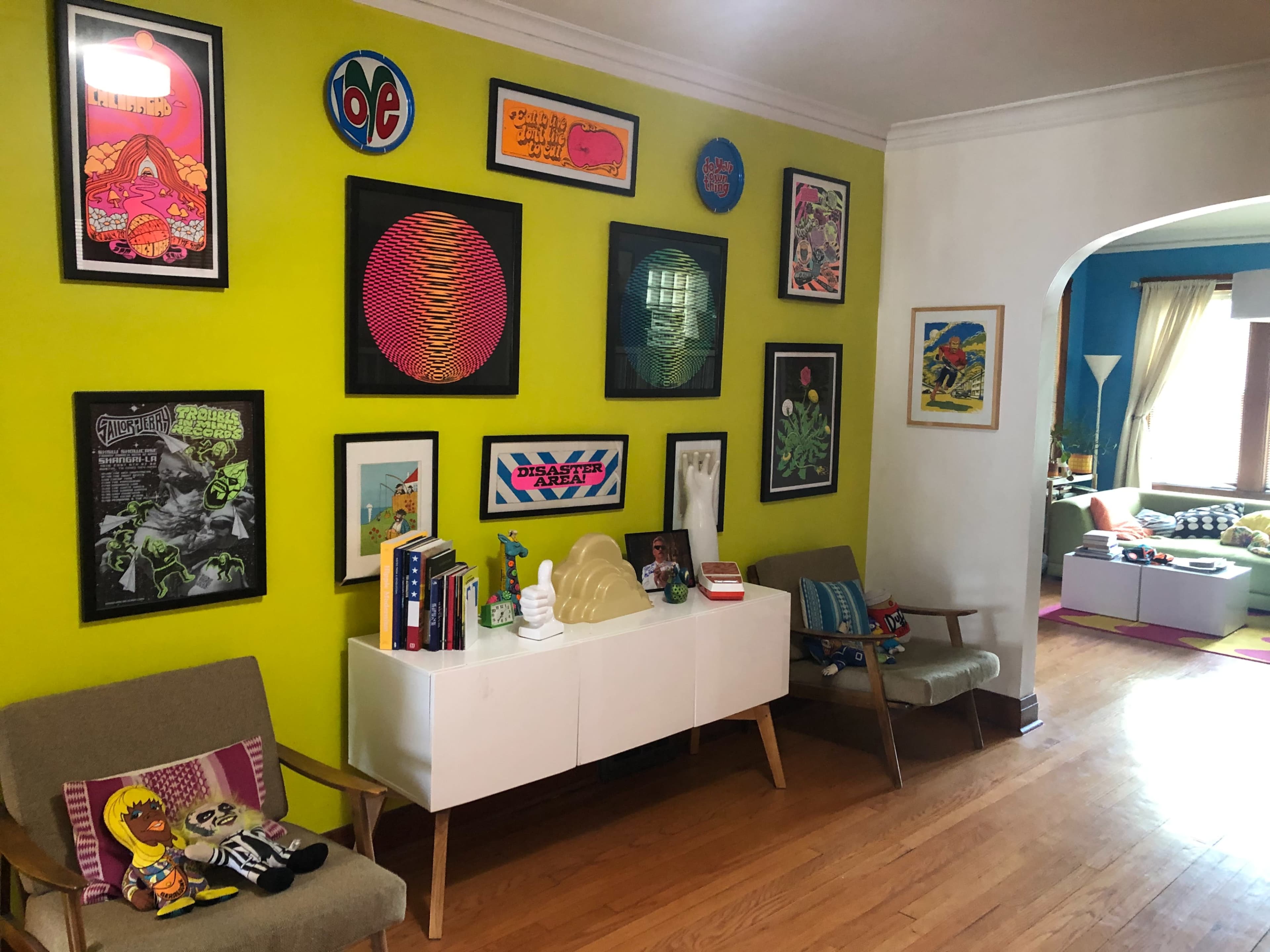 The image showcases a brightly colored living room with a green accent wall adorned with various framed artworks, including prints and posters, while a white sideboard holds books and decorative items.