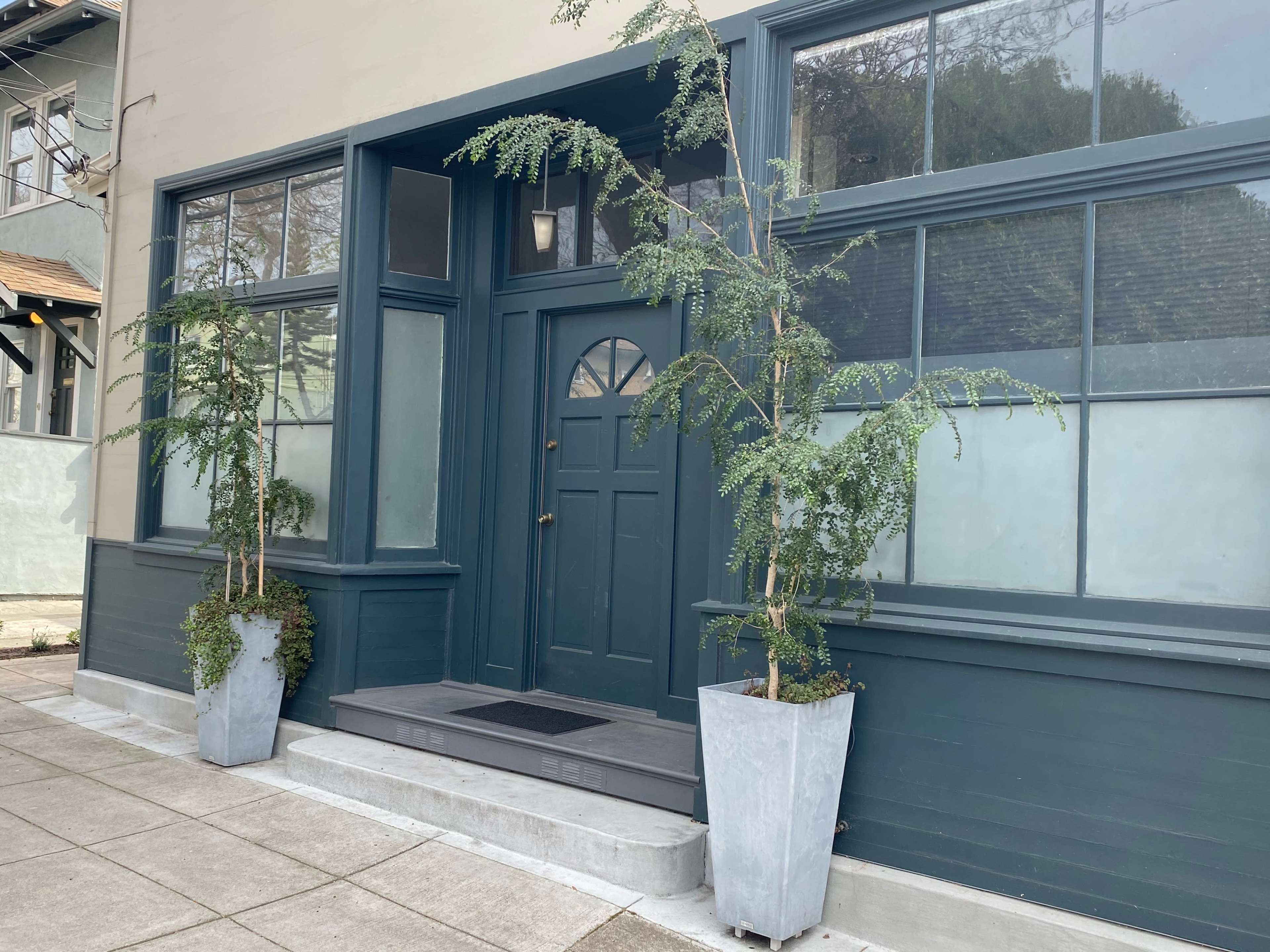 A modern, blue-gray facade of a building with a central door flanked by two tall planters containing plants.