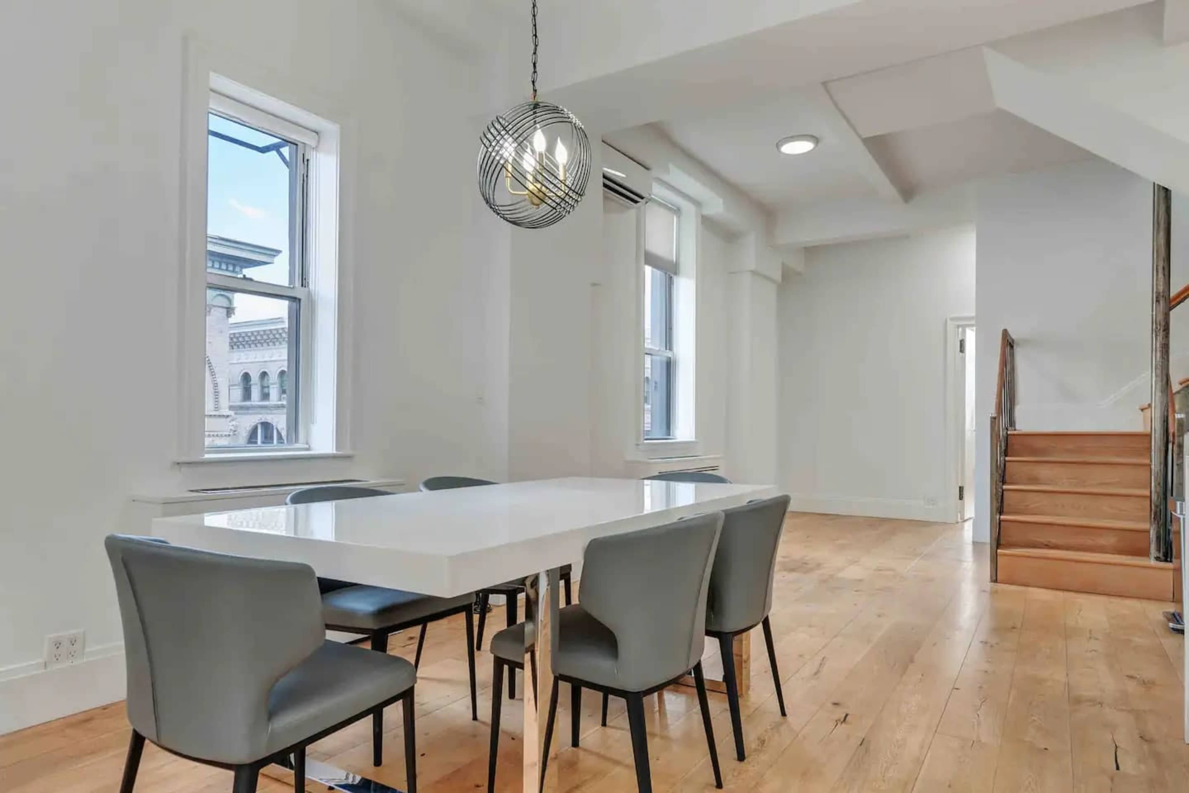 A modern dining area with a white table and gray chairs, featuring large windows and a wooden staircase in the background.