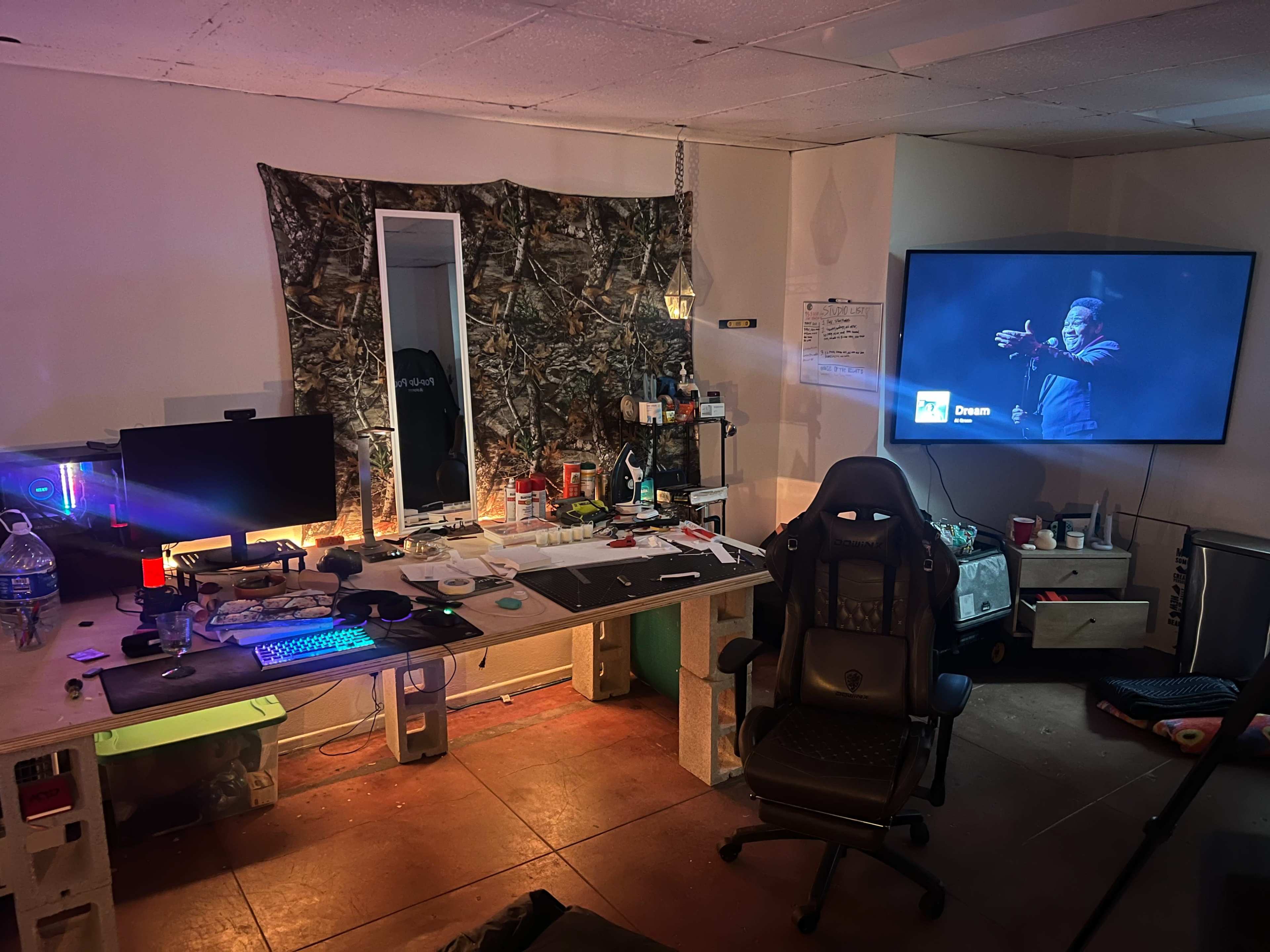 The image depicts a cluttered room featuring a desk with a computer monitor, a gaming chair, a large television mounted on the wall, and various items scattered across the workspace.