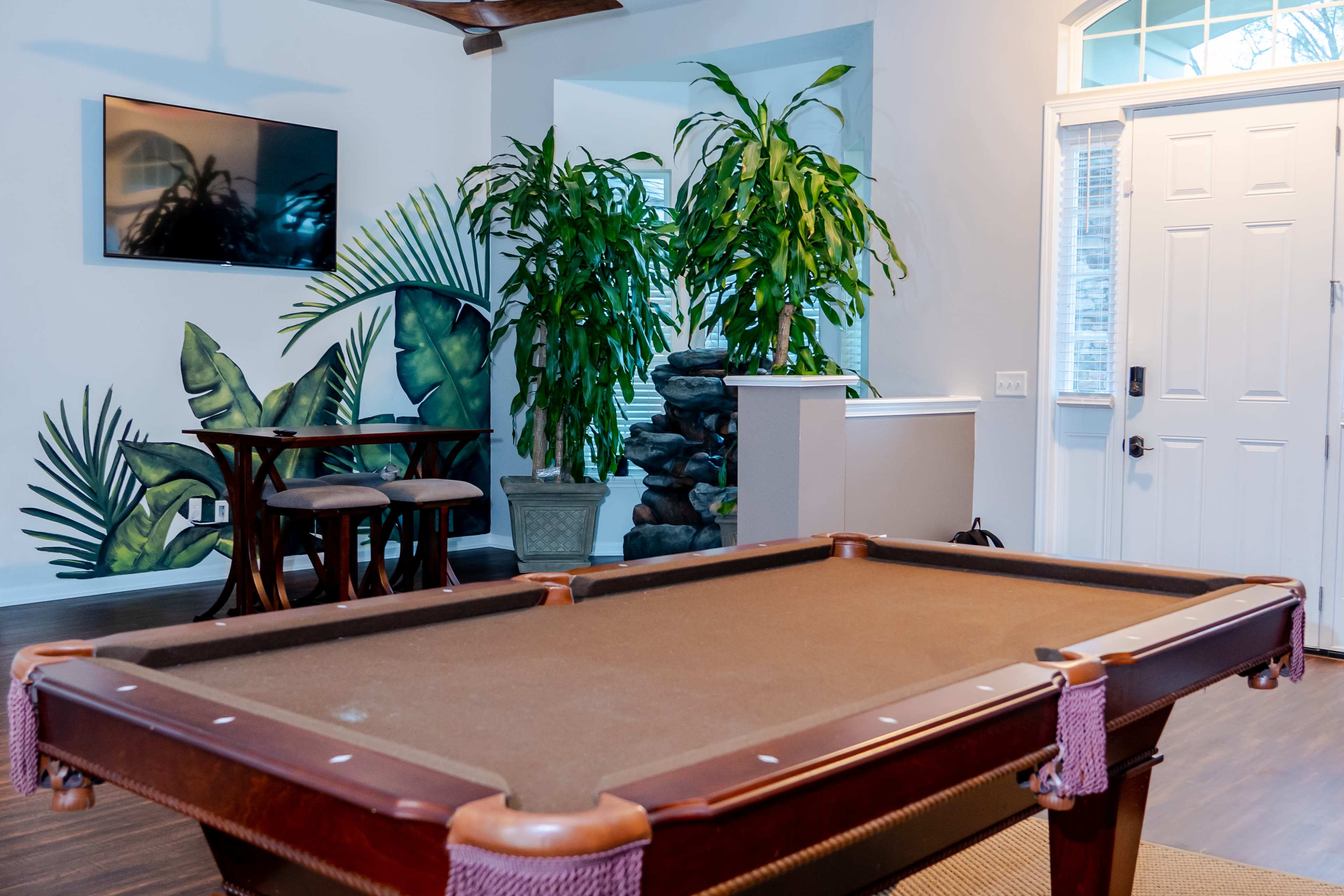 A pool table in a room with a television on the wall, large plants, and a doorway leading outside.
