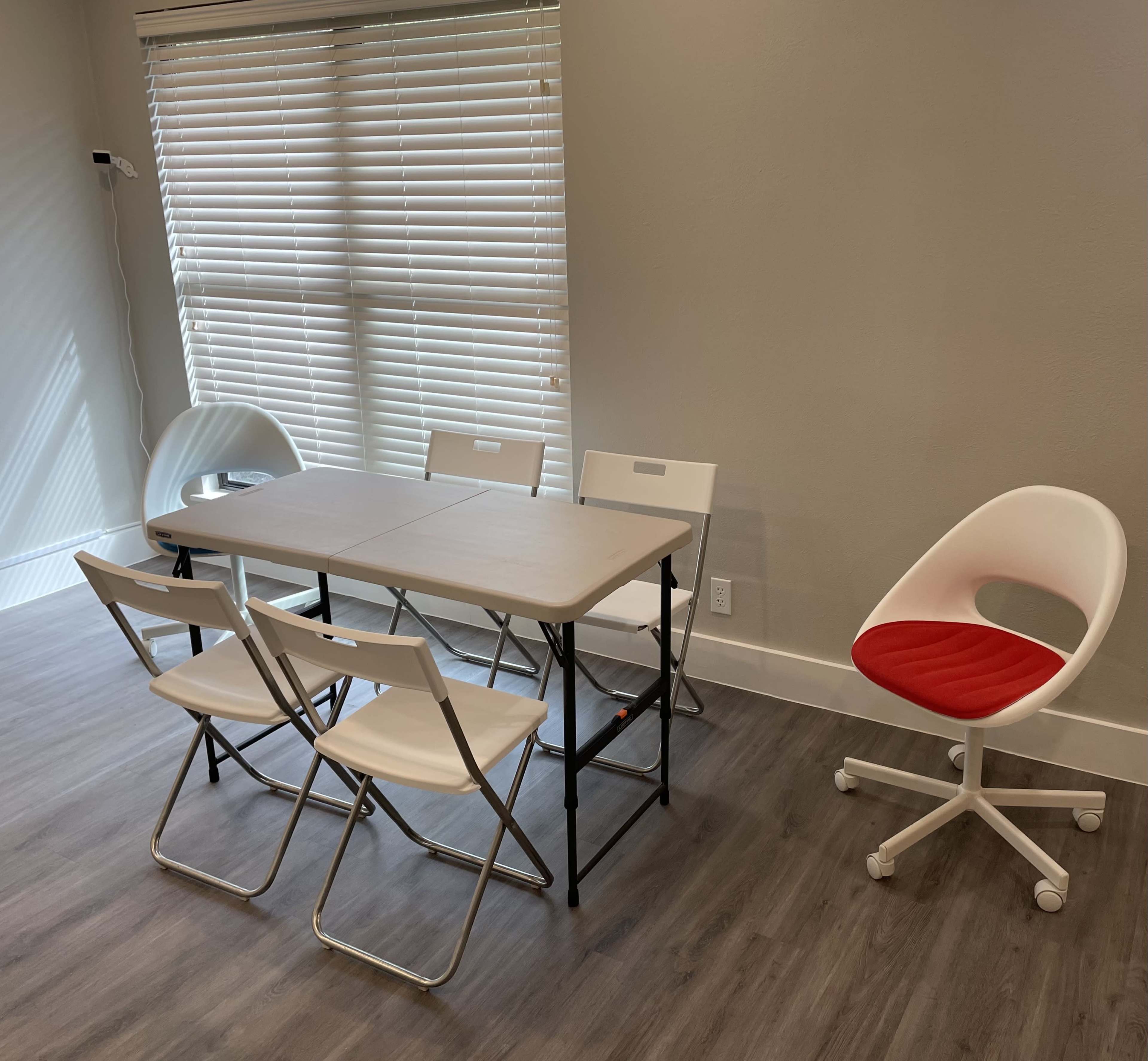 A small room features a rectangular table surrounded by four folding chairs, with a red-accented office chair positioned nearby.