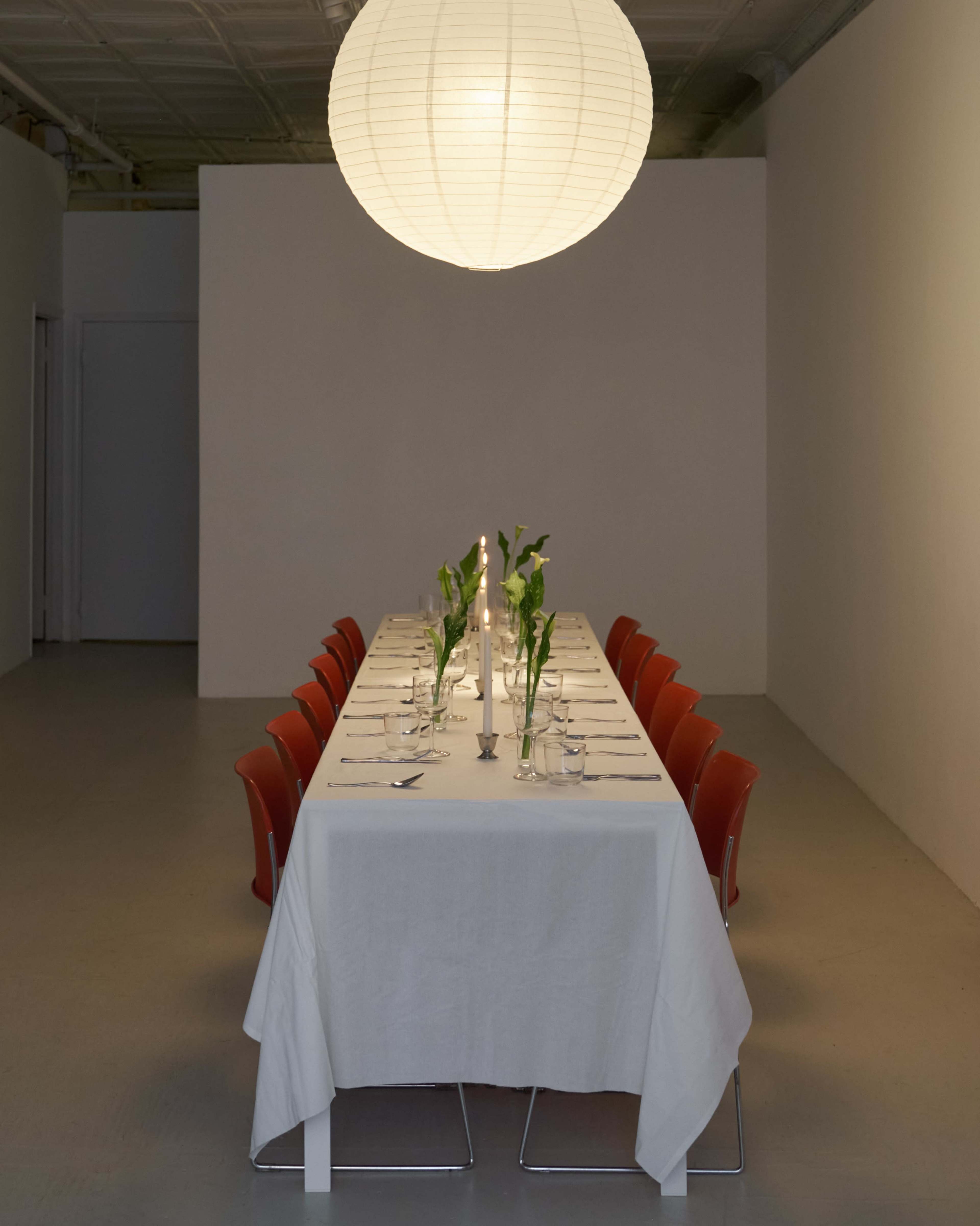 A long dining table is set with white tableware and decorated with green plants, illuminated by a large paper lantern overhead.