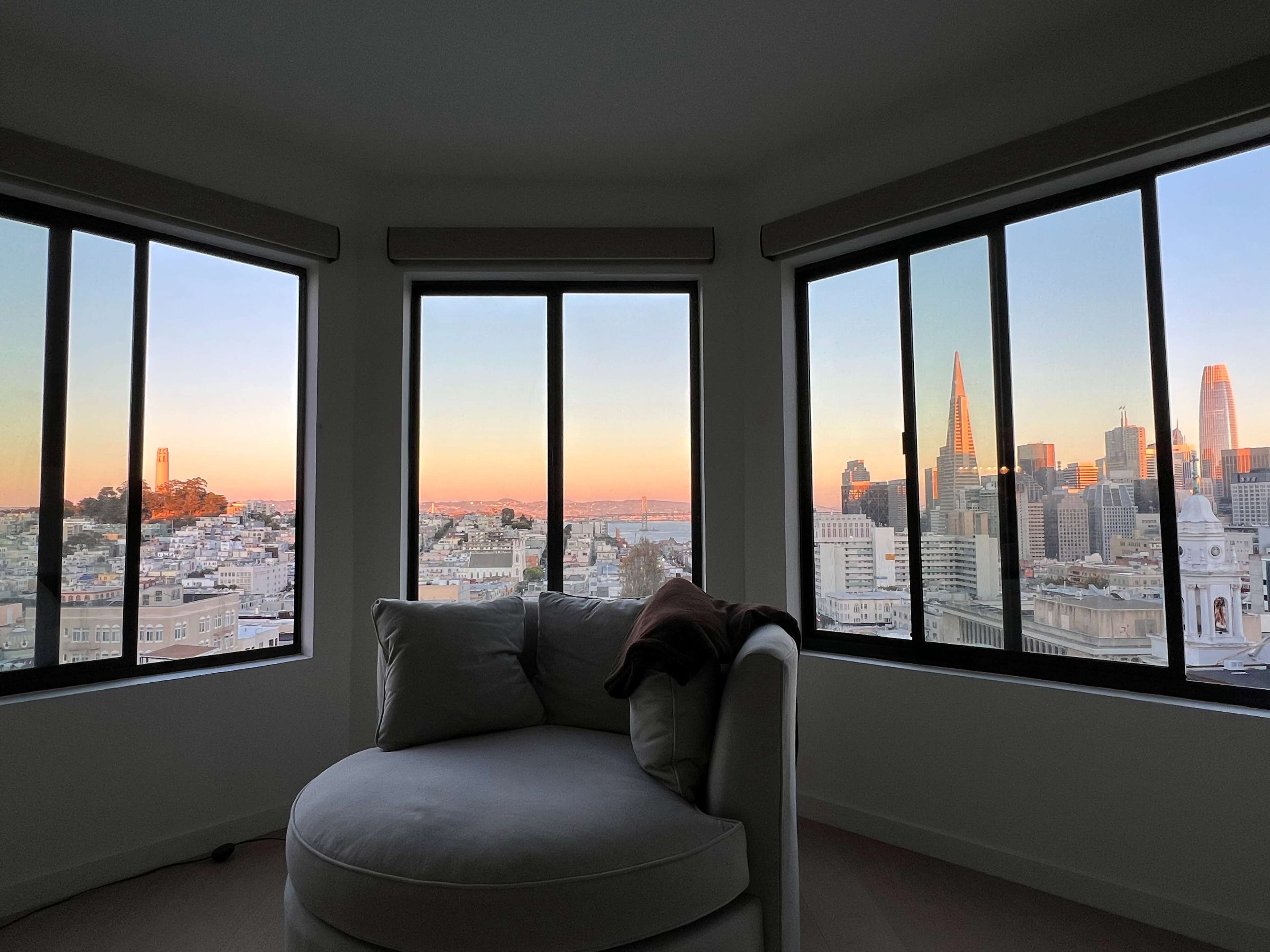 A cozy corner with a circular sofa, framed by large windows that offer a view of the San Francisco skyline during sunset.