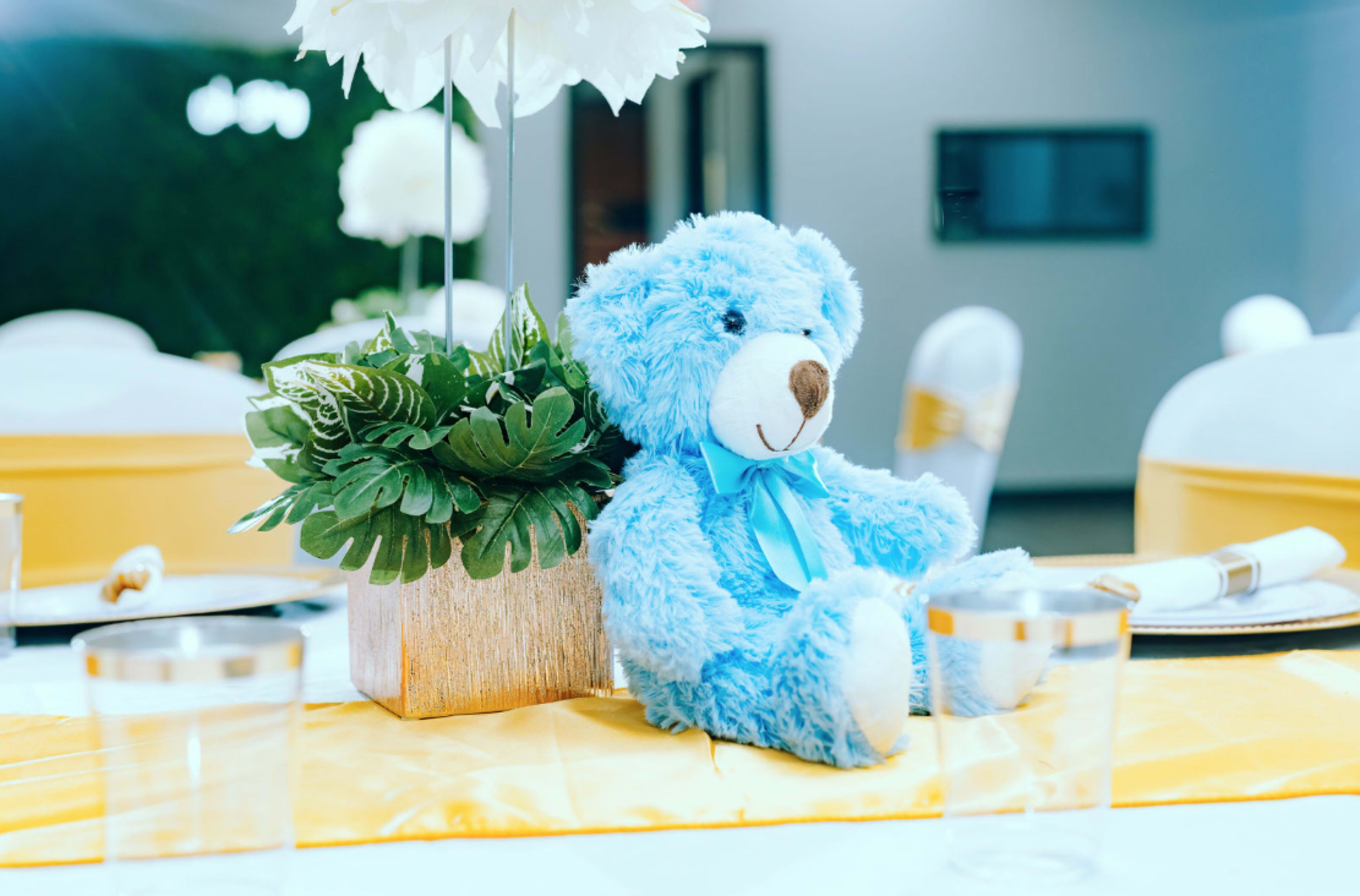 A blue teddy bear is seated next to a floral centerpiece on a table set for an event.