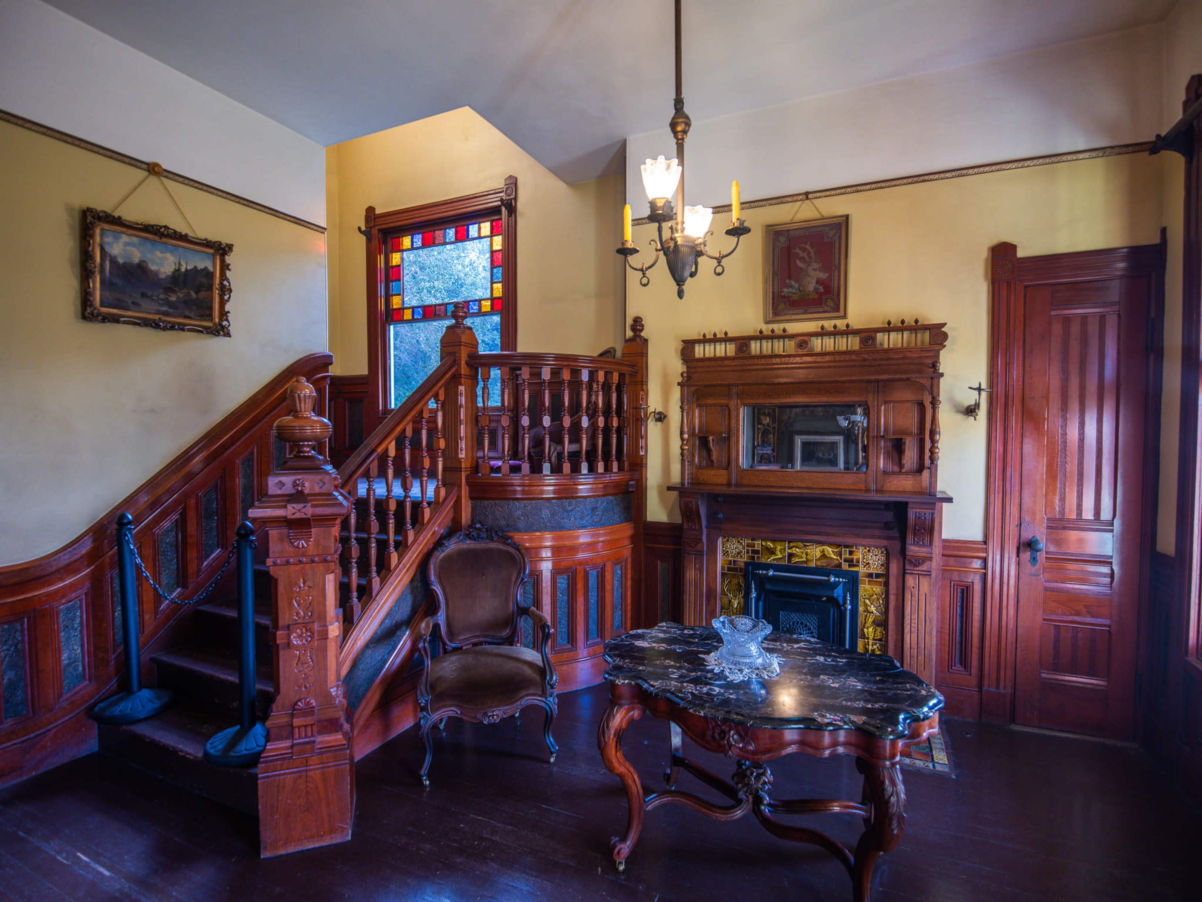 A vintage interior featuring a wooden staircase, a decorative fireplace, and a dark table in a room with stained glass windows.