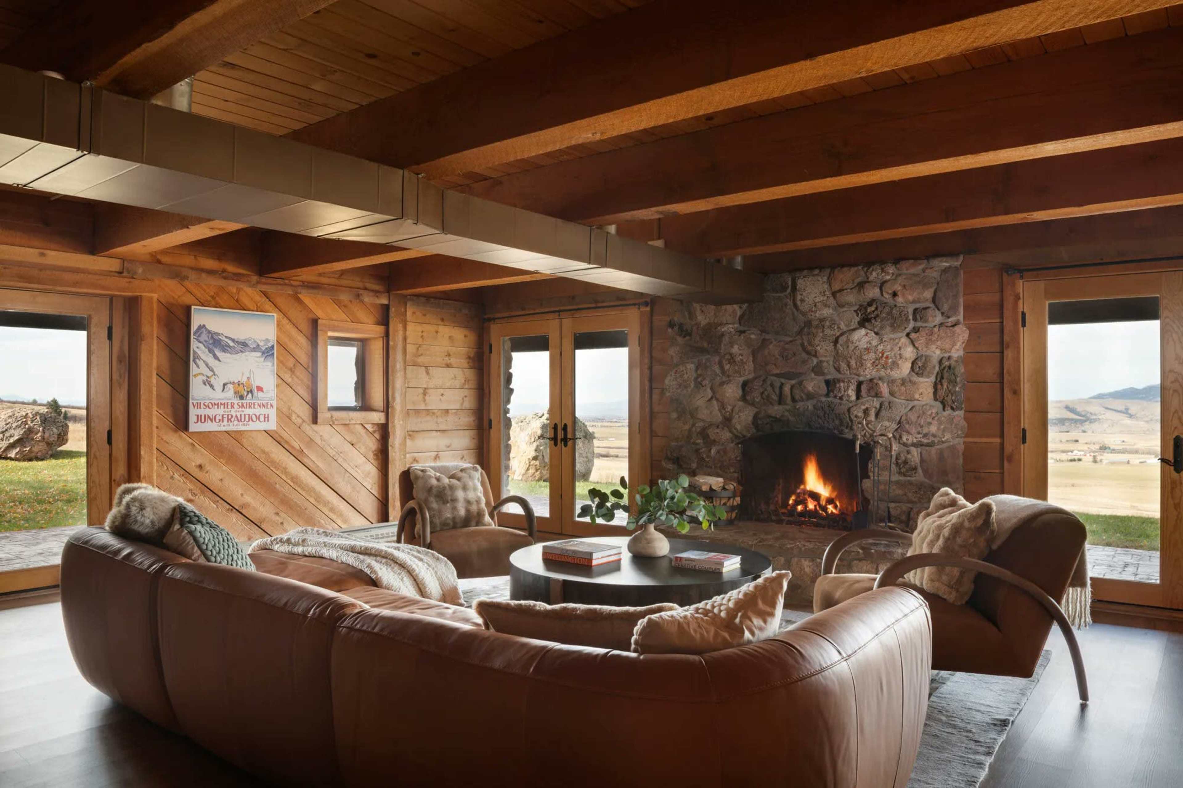 The image shows a cozy living room featuring a circular leather sofa, a stone fireplace, and large glass doors that provide scenic views of the outdoors.