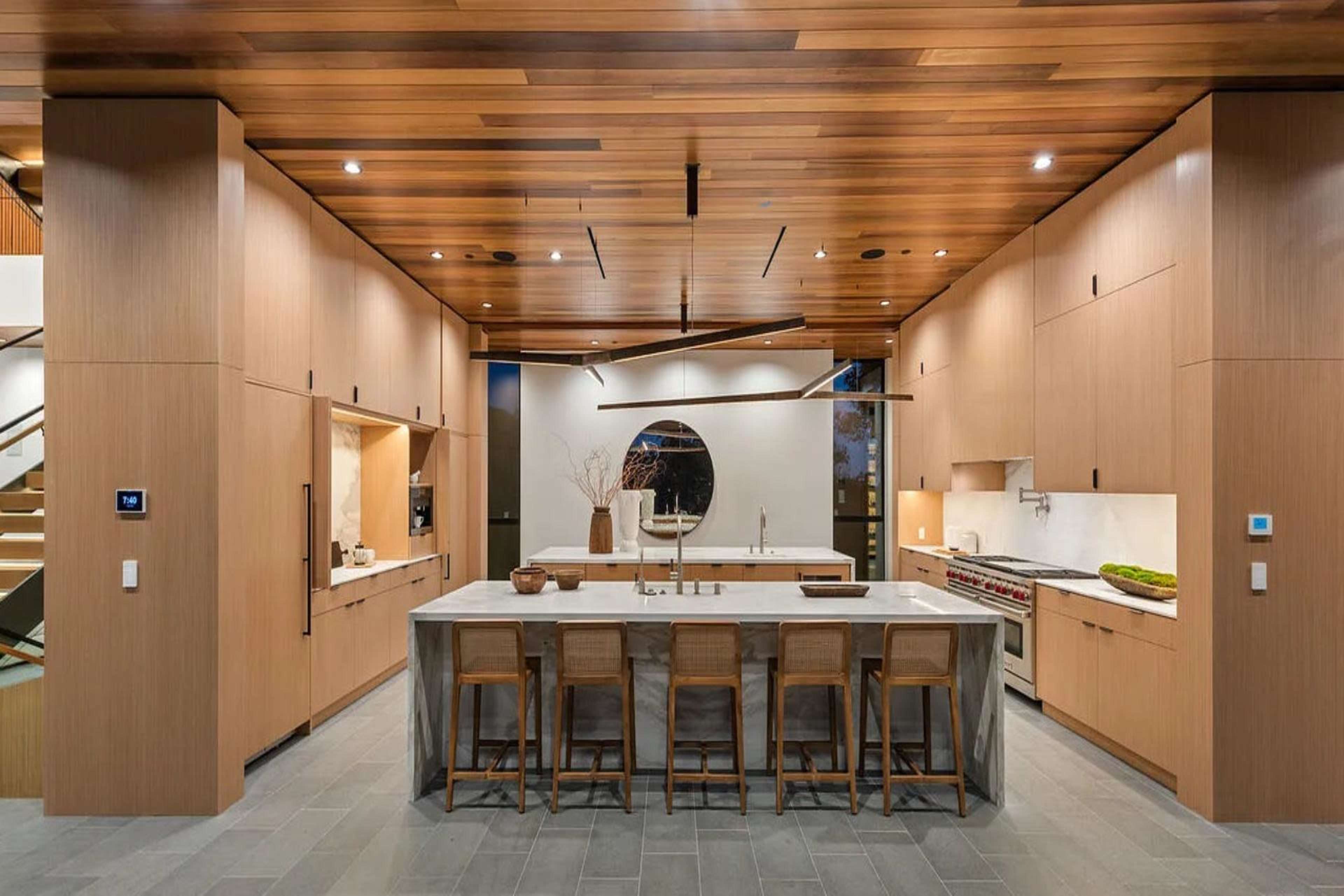 The image shows a modern kitchen with a large marble island, wooden cabinetry, and a row of bar stools.