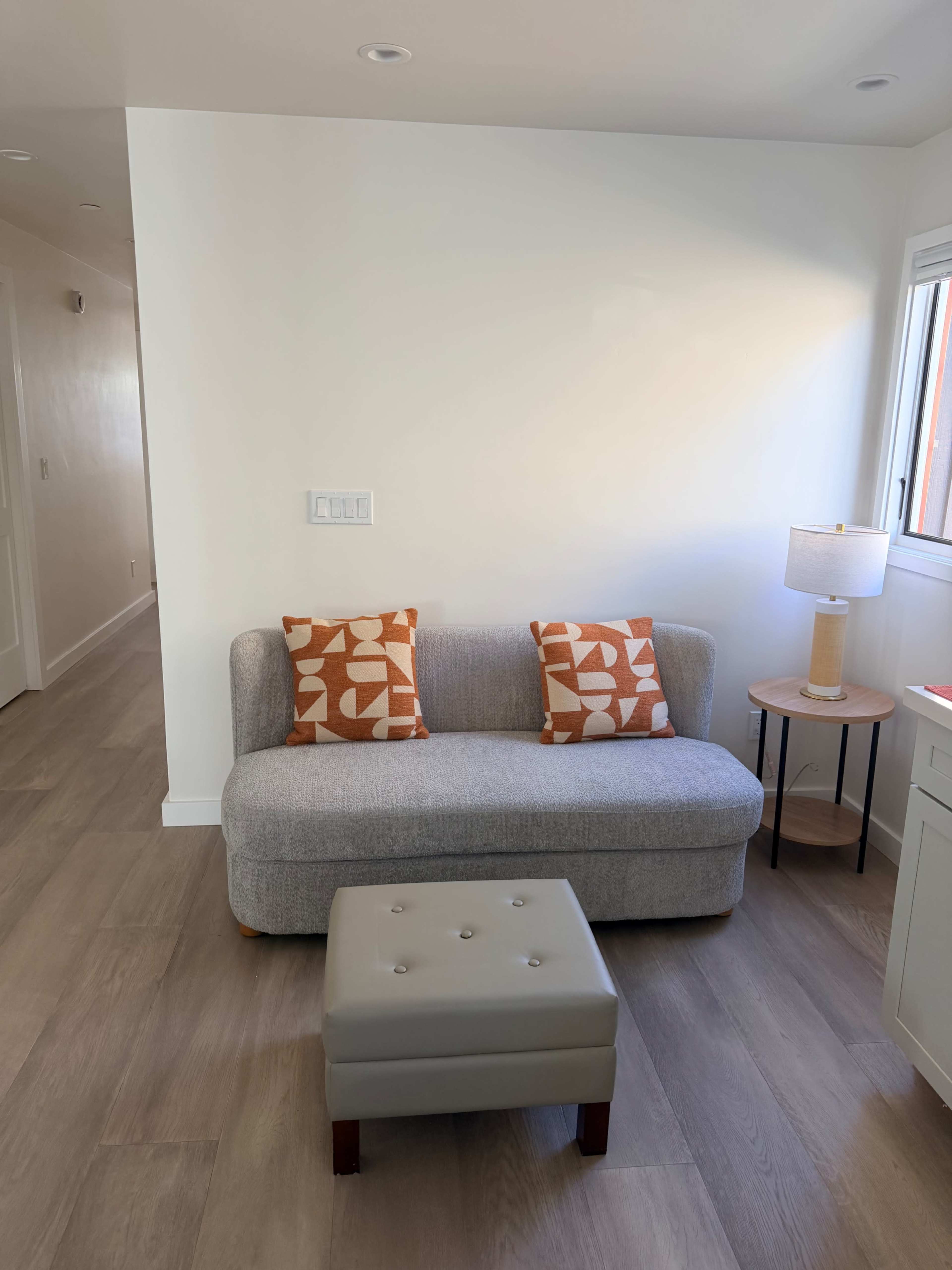 A small room features a gray couch with patterned orange pillows, a lamp on a side table, and a tufted ottoman on a wooden floor.