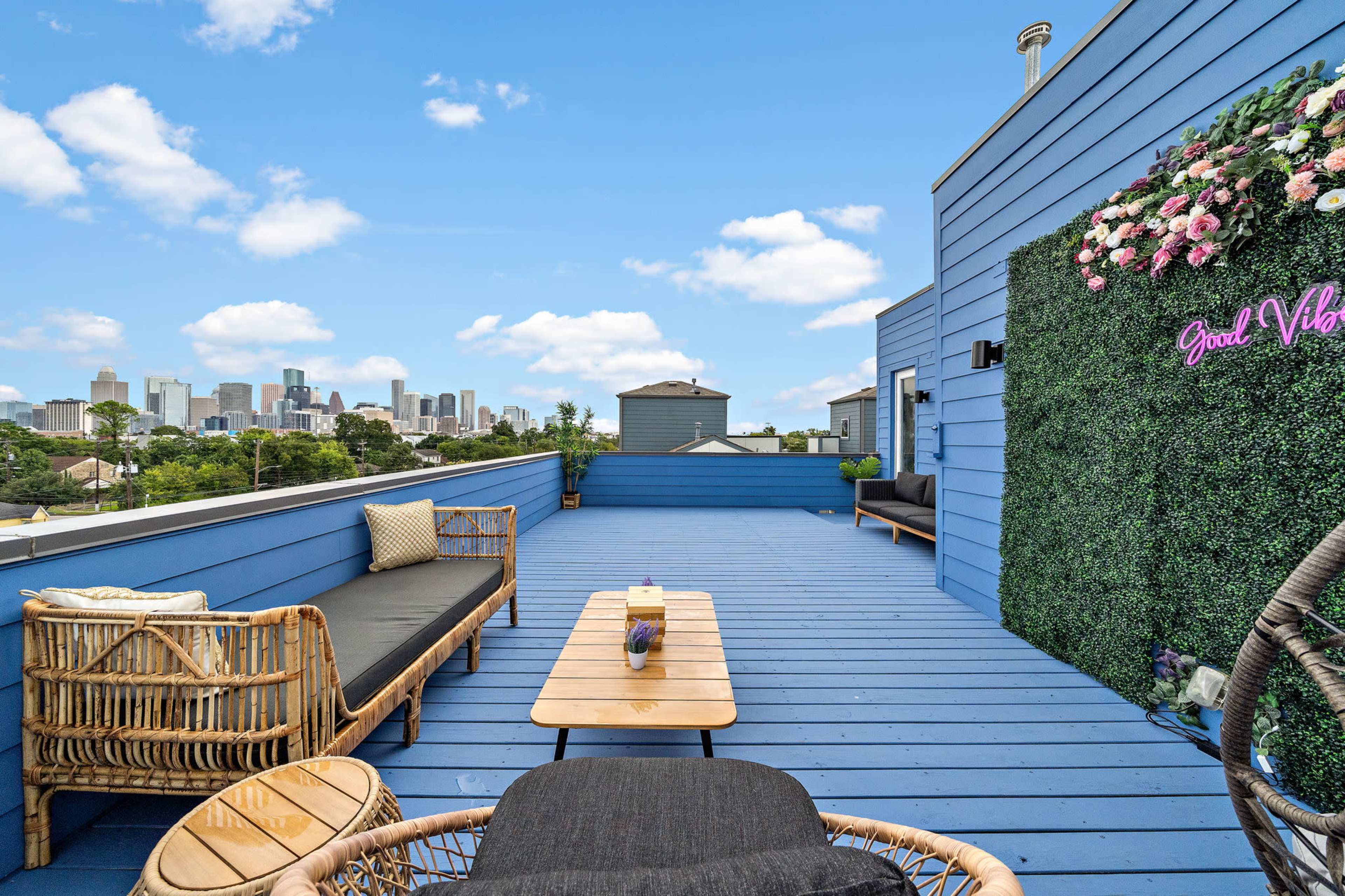 A rooftop patio features wicker furniture and a floral wall, offering a view of a city skyline under a clear blue sky.