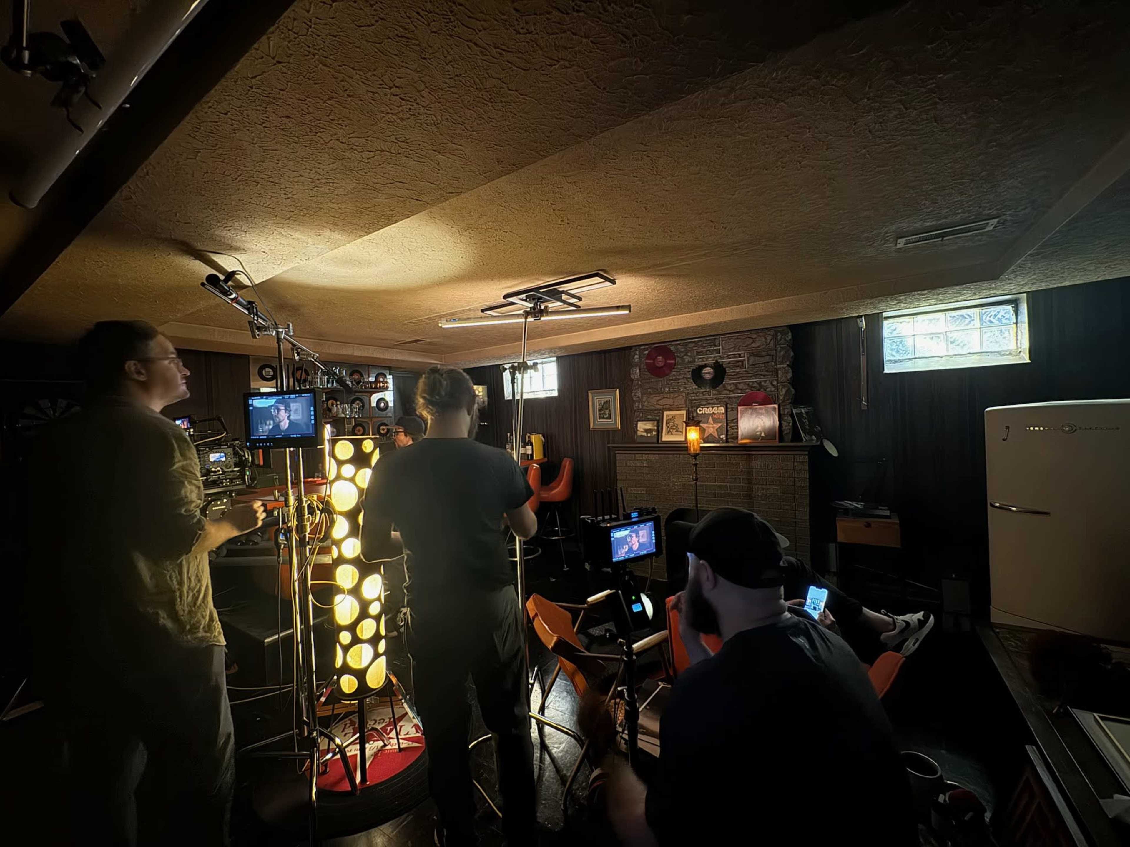 Three people operate filming equipment in a dimly lit basement space filled with various items and set pieces.