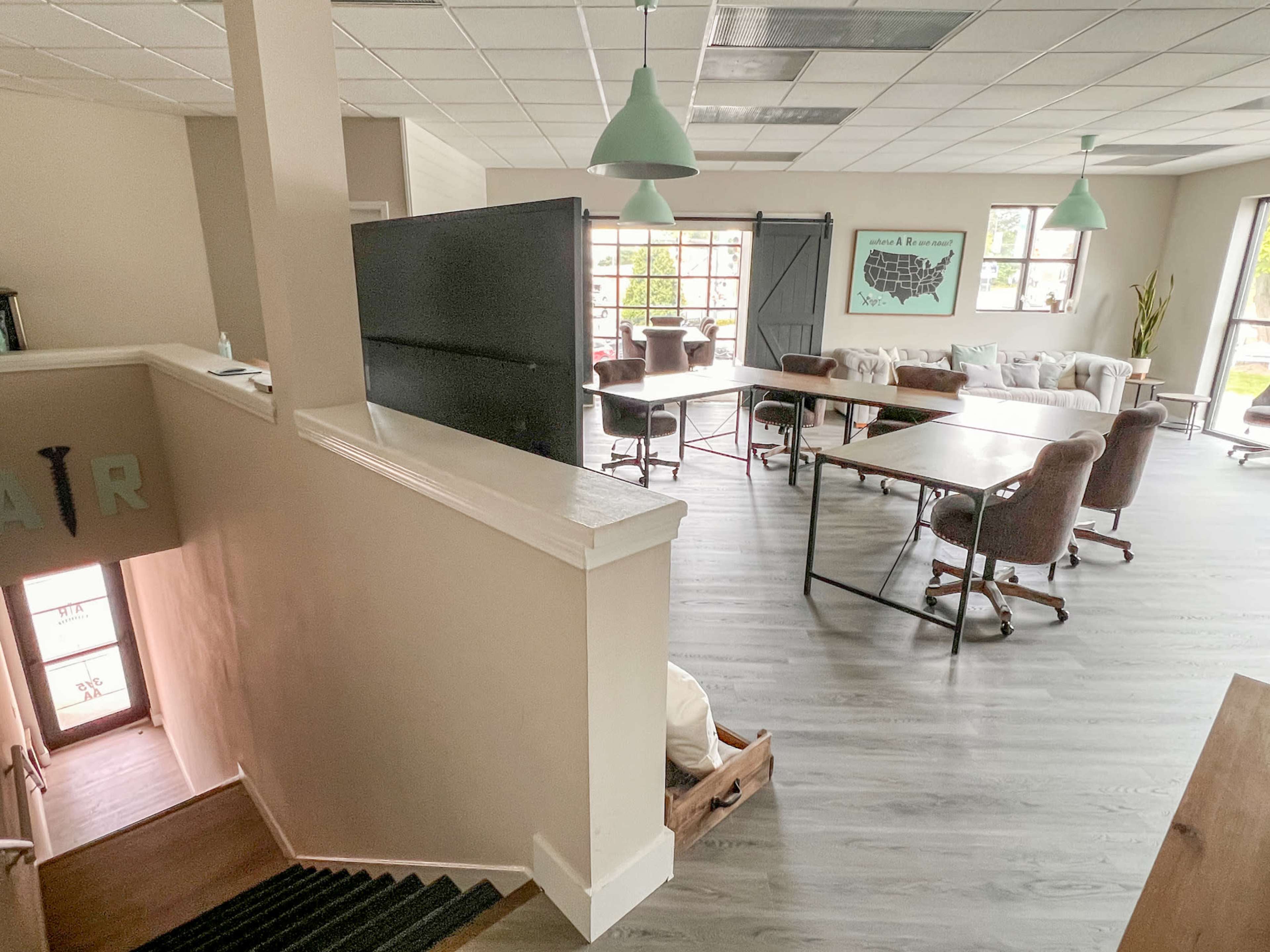 The image shows a well-lit, open workspace featuring several desks, chairs, and a staircase leading to the entrance.