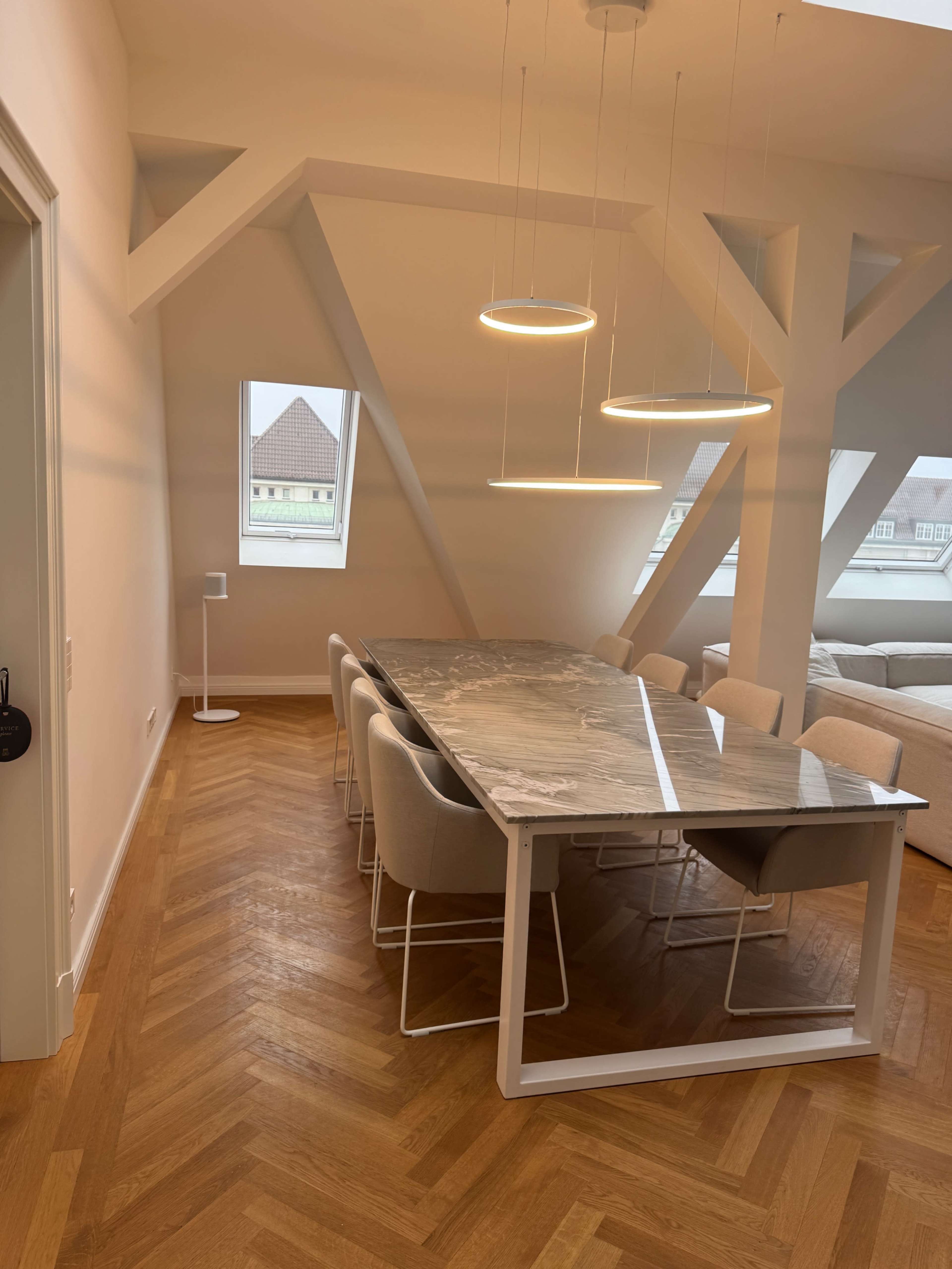 The image shows a modern dining area with a long marble table surrounded by upholstered chairs, illuminated by circular pendant lights, set under a sloped ceiling.