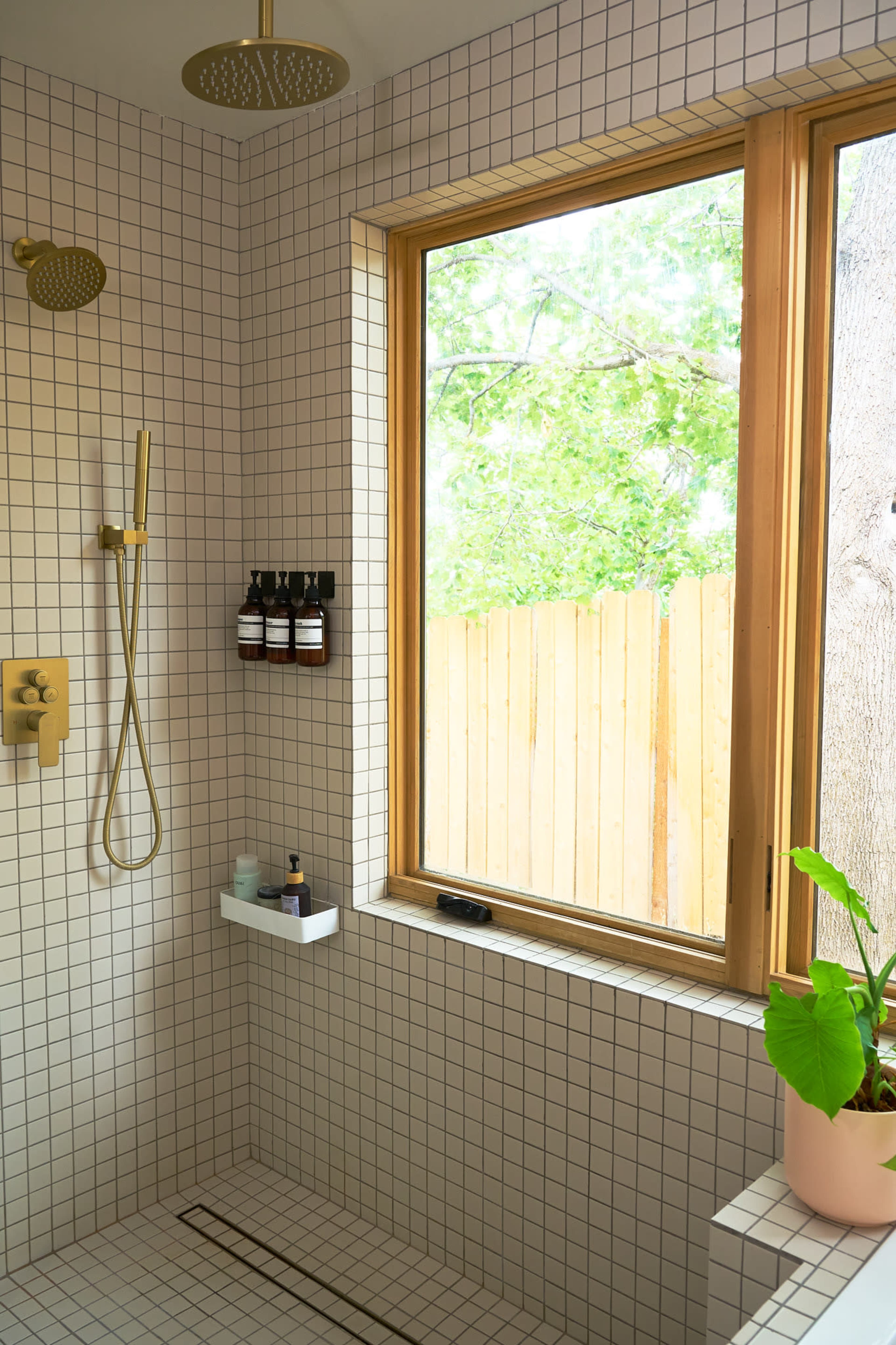 The image shows a tiled shower area with a window overlooking greenery, featuring a showerhead, bottles on a shelf, and a potted plant beside the window.