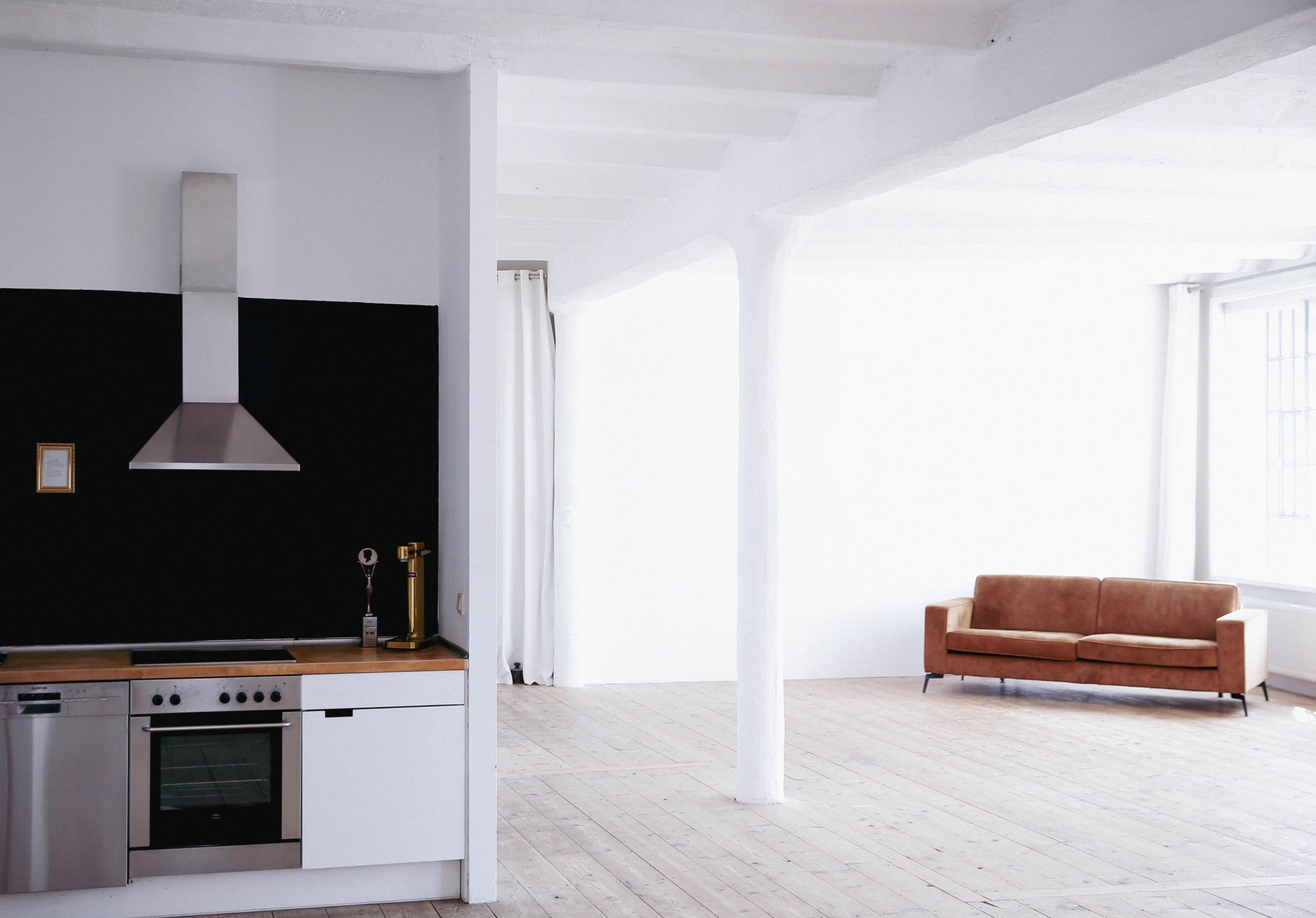 A modern kitchen with stainless steel appliances is adjacent to an empty living area featuring a brown sofa against a white wall.