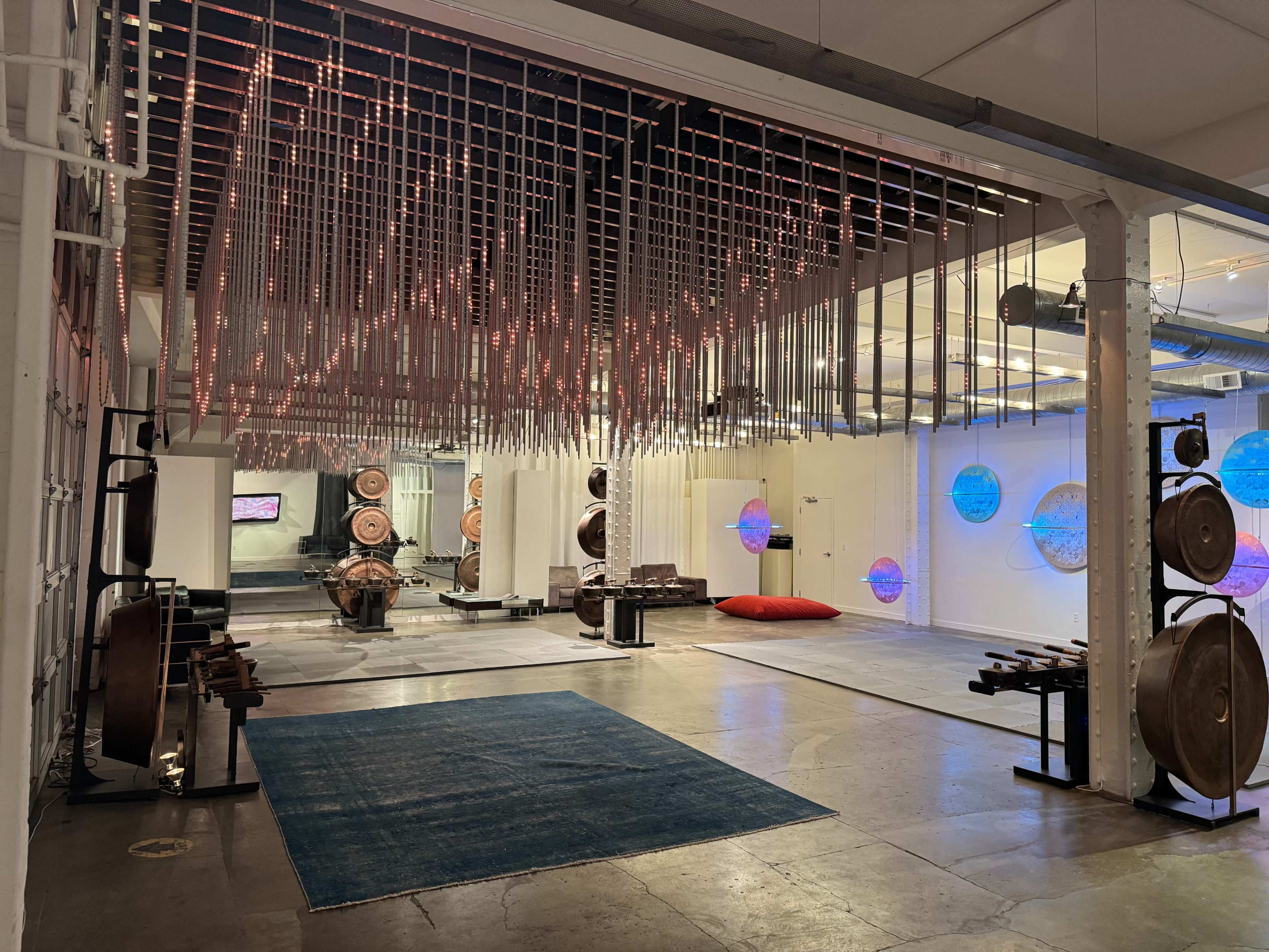 The image shows a modern, industrial space with a ceiling adorned with hanging metal rods, an open floor area featuring various musical instruments, and a large red cushion on the ground.