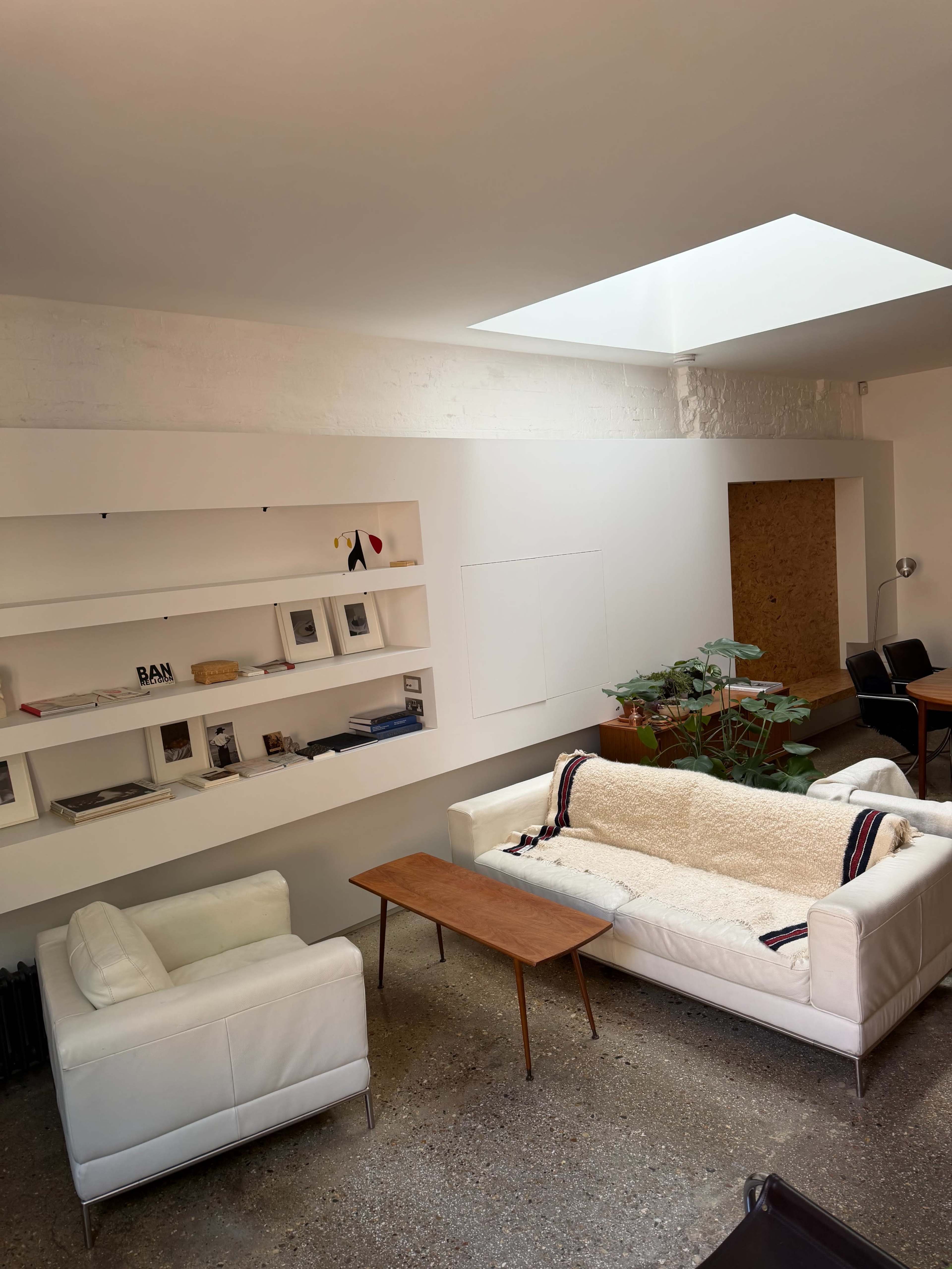A minimalist living room features two white sofas, a wooden coffee table, and a skylight, with shelves displaying books and decorative items along the wall.