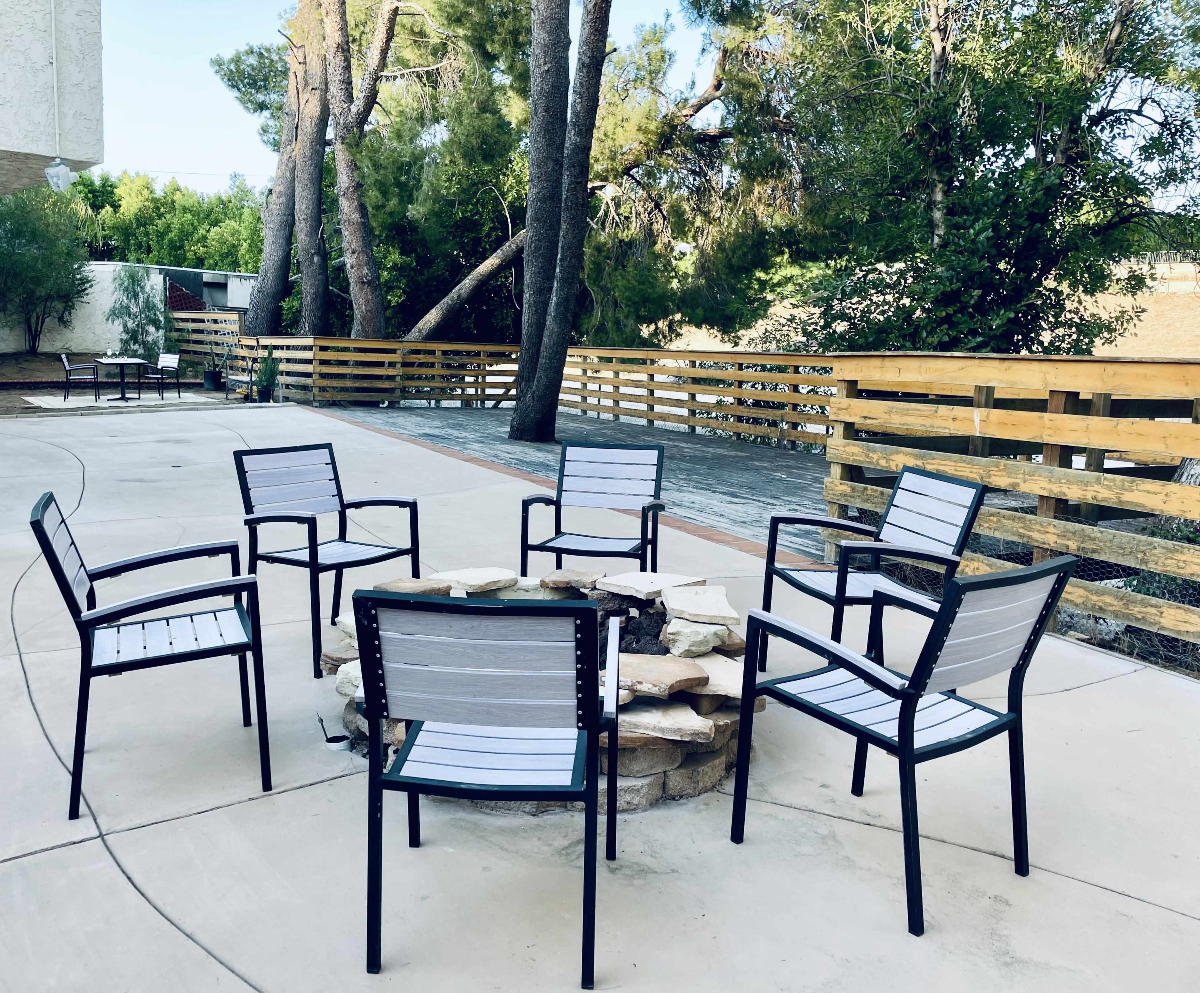 A circular stone fire pit is surrounded by six metal chairs on a paved outdoor area, with trees and wooden fencing in the background.
