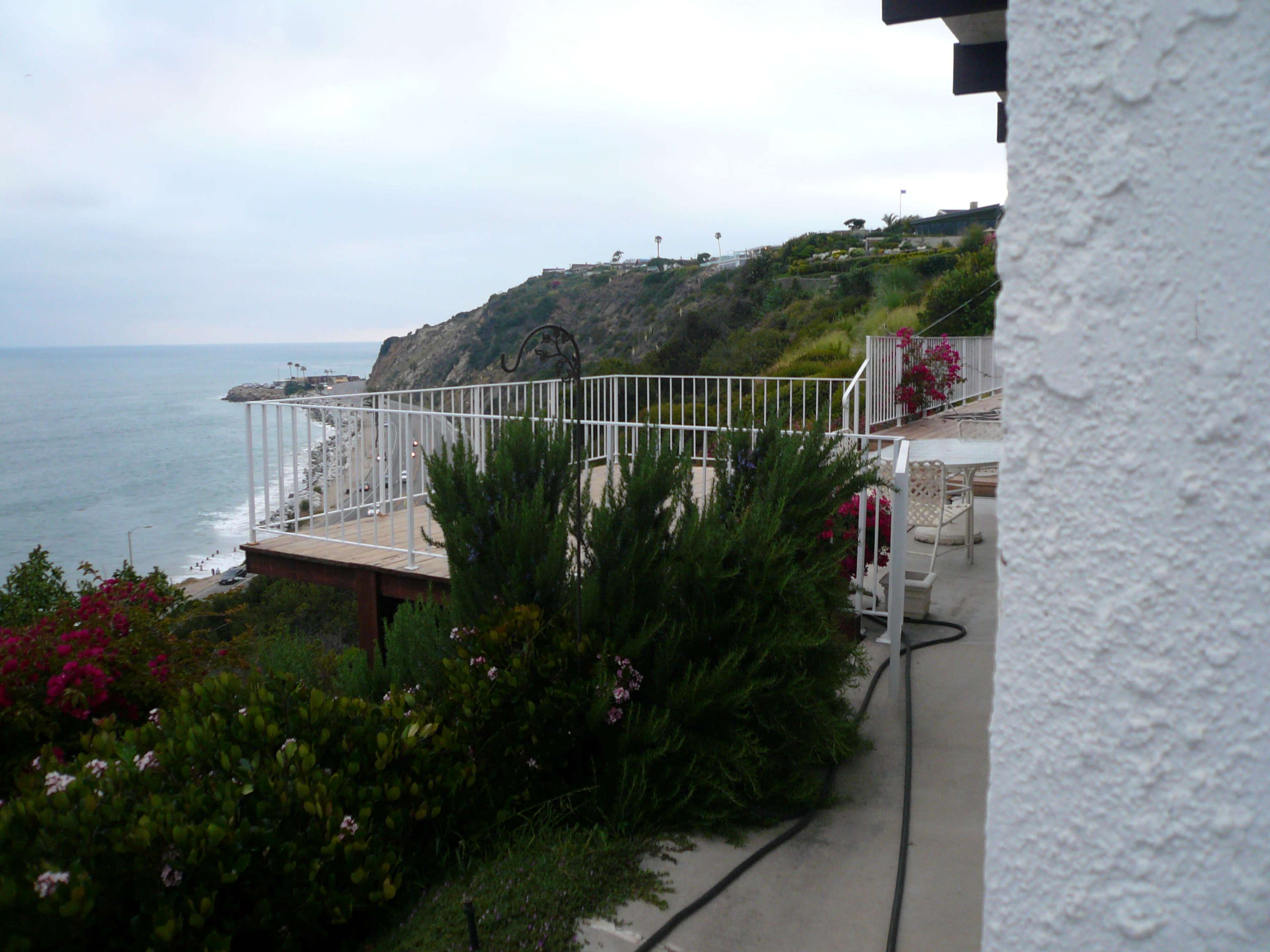 The image shows a coastal view featuring a deck overlooking the ocean, surrounded by greenery and flowering plants.