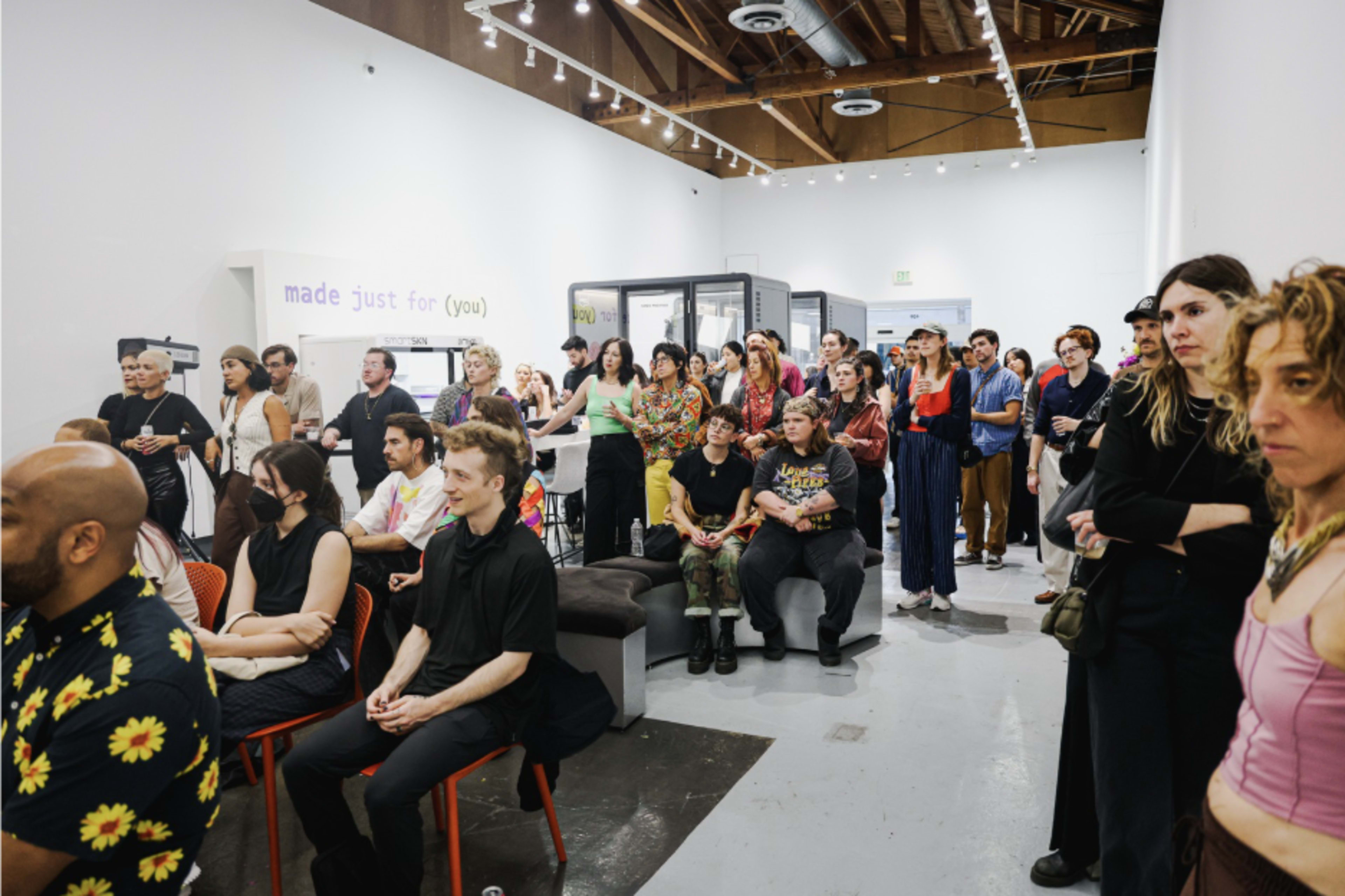 A crowd of diverse individuals is gathered in an art gallery for an event, with some seated and others standing.