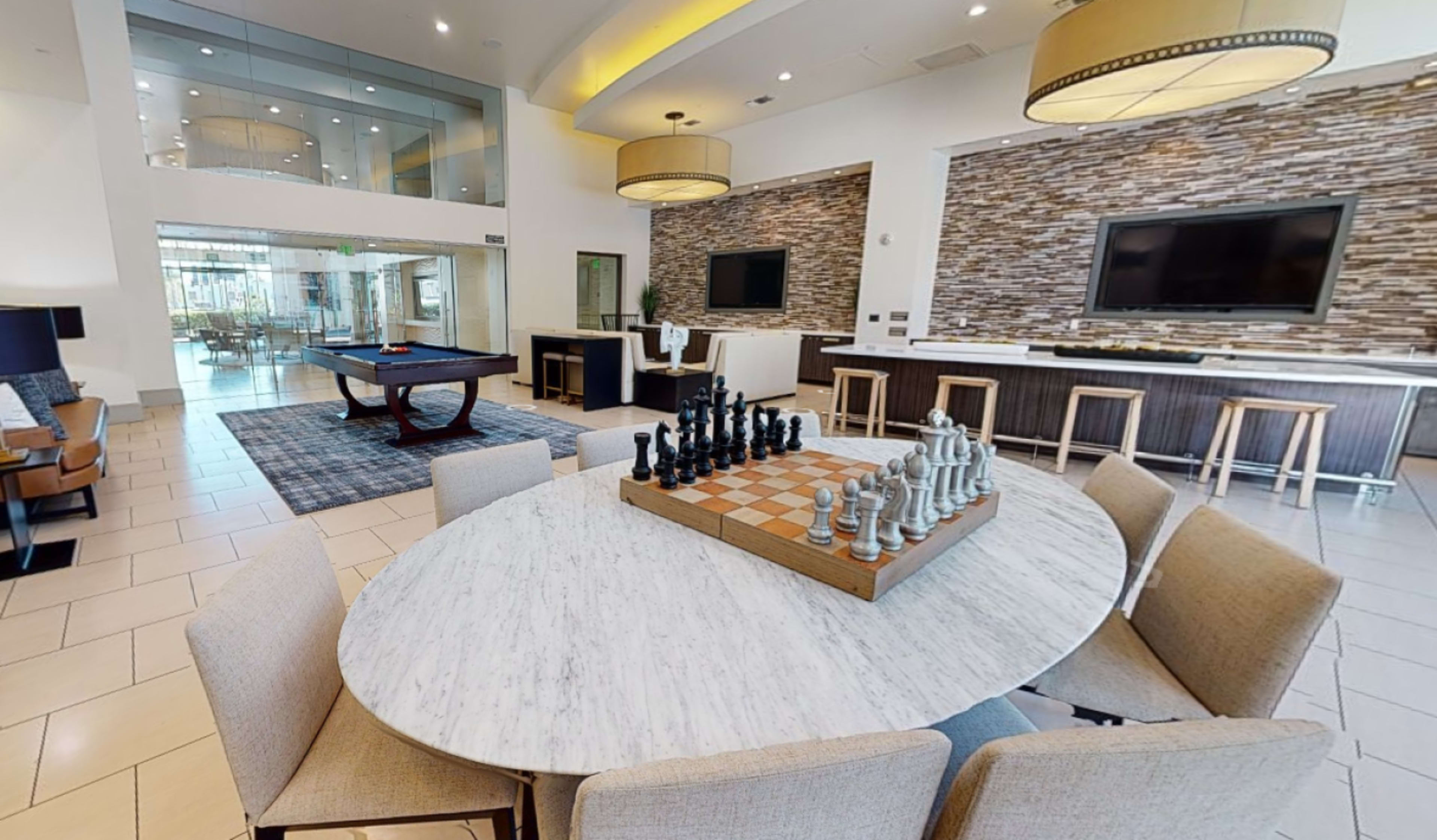 A modern communal lounge features a large round table with a chess set, seating area, and a game room visible in the background.