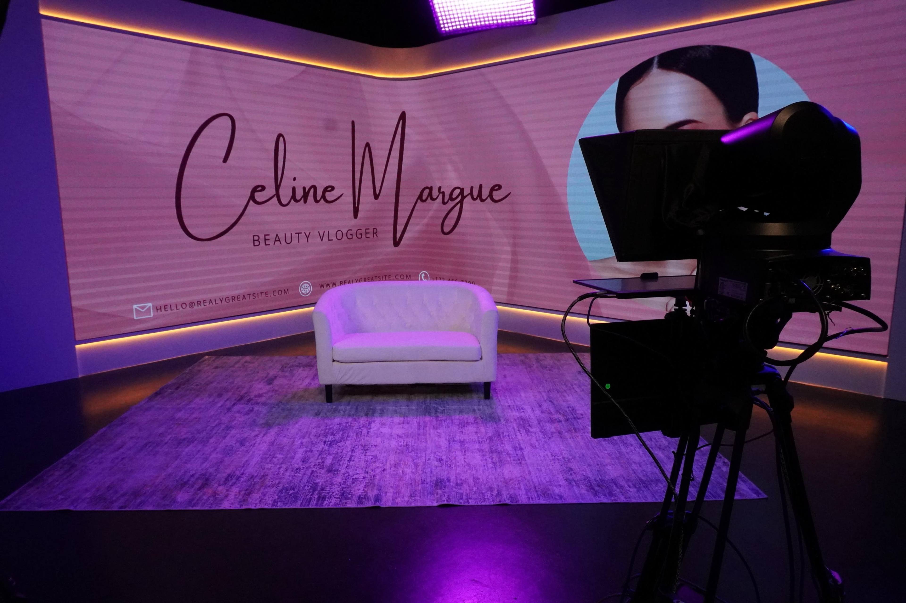 The image shows a television studio setting with a white couch and a backdrop featuring the name "Celine Margue, Beauty Vlogger," along with a camera pointed towards the seating area.