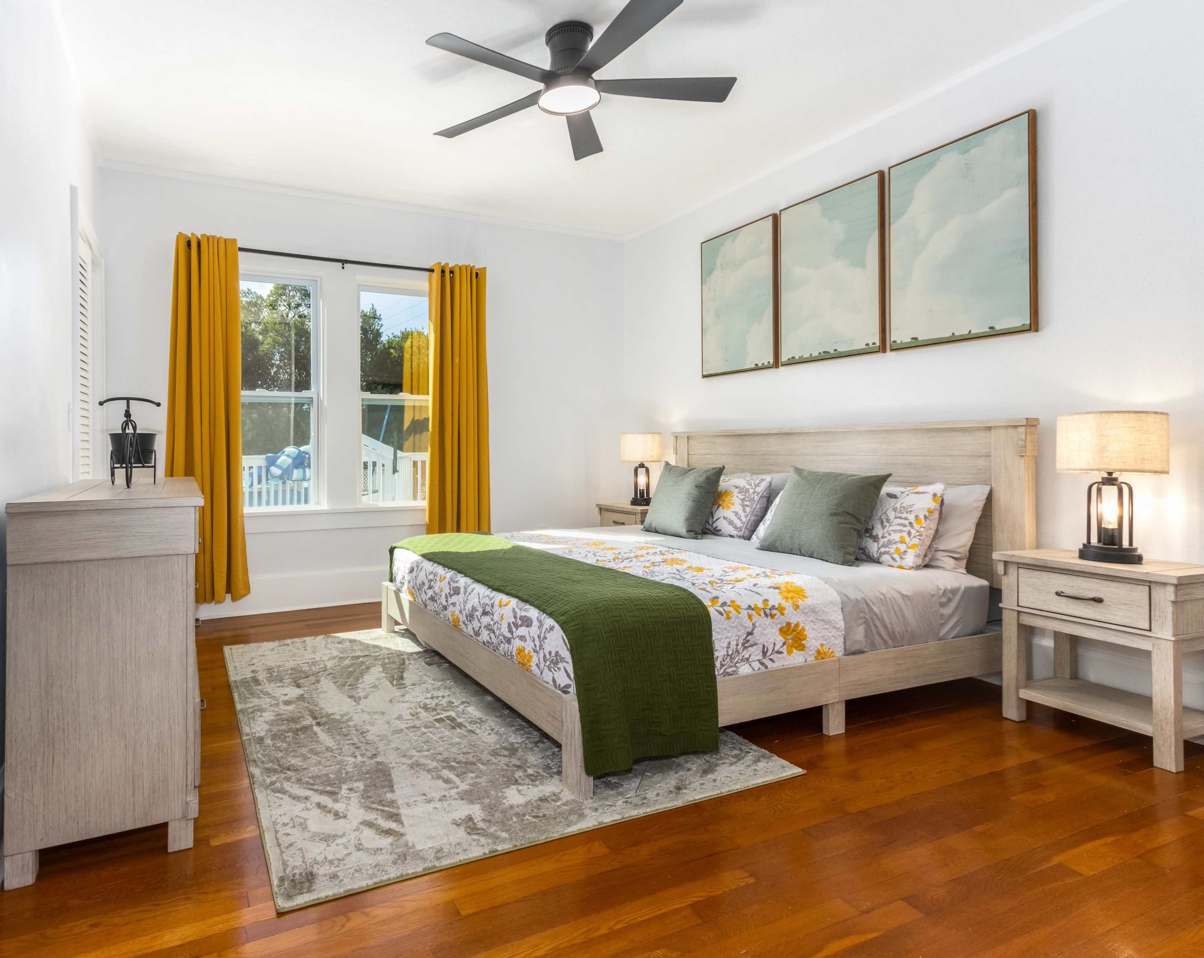 The bedroom features a large bed with floral bedding, two nightstands with lamps, a dresser, and large windows with yellow curtains.