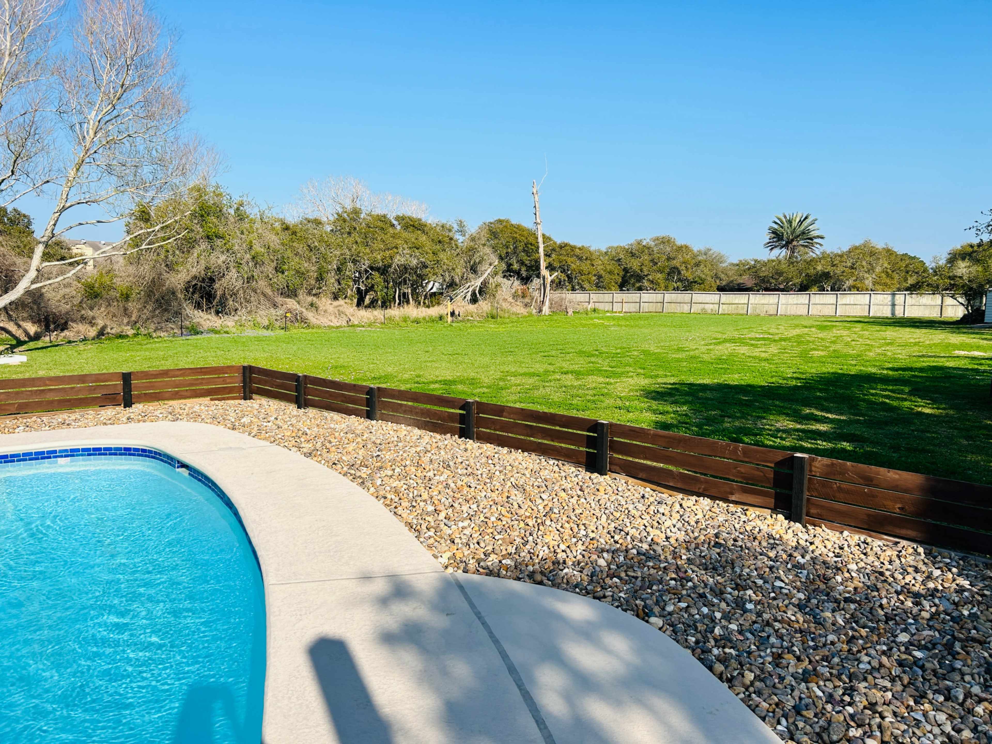 A swimming pool with a stone border is adjacent to a manicured grassy area surrounded by trees and a wooden fence.