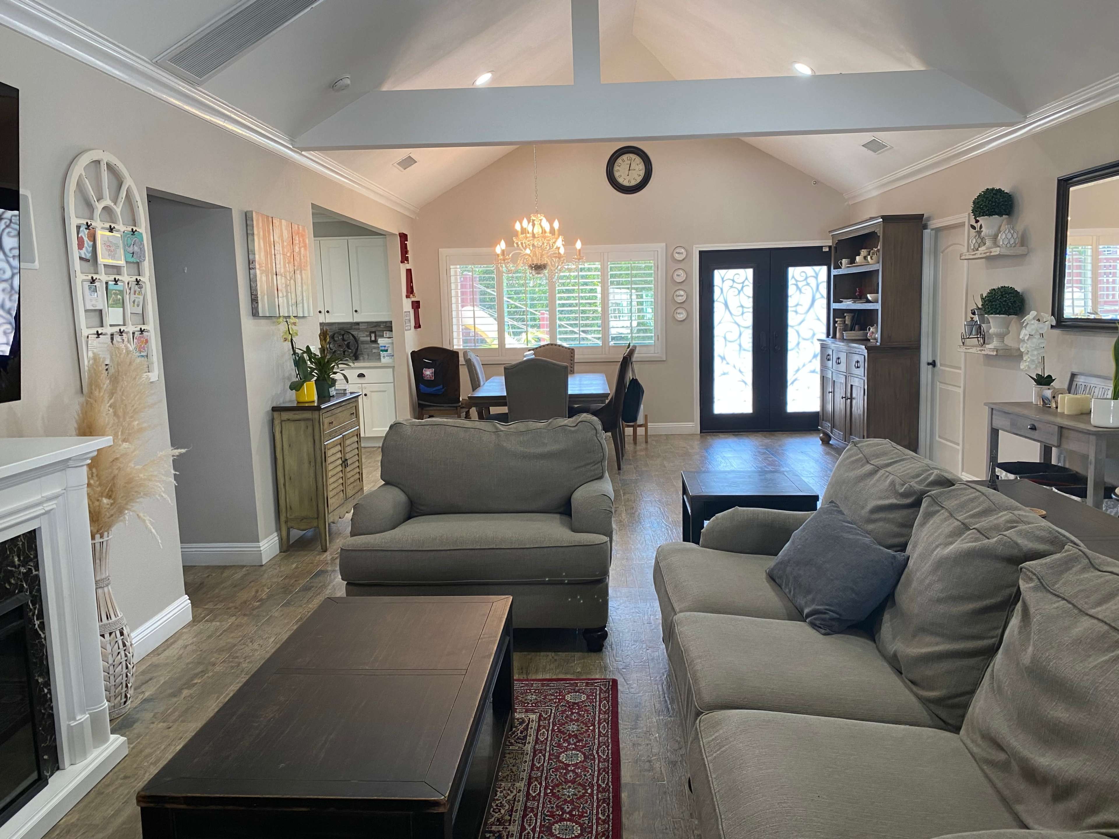 A living room features a gray sofa and coffee table, with an adjacent dining area visible through a set of double doors, and light wood flooring throughout.