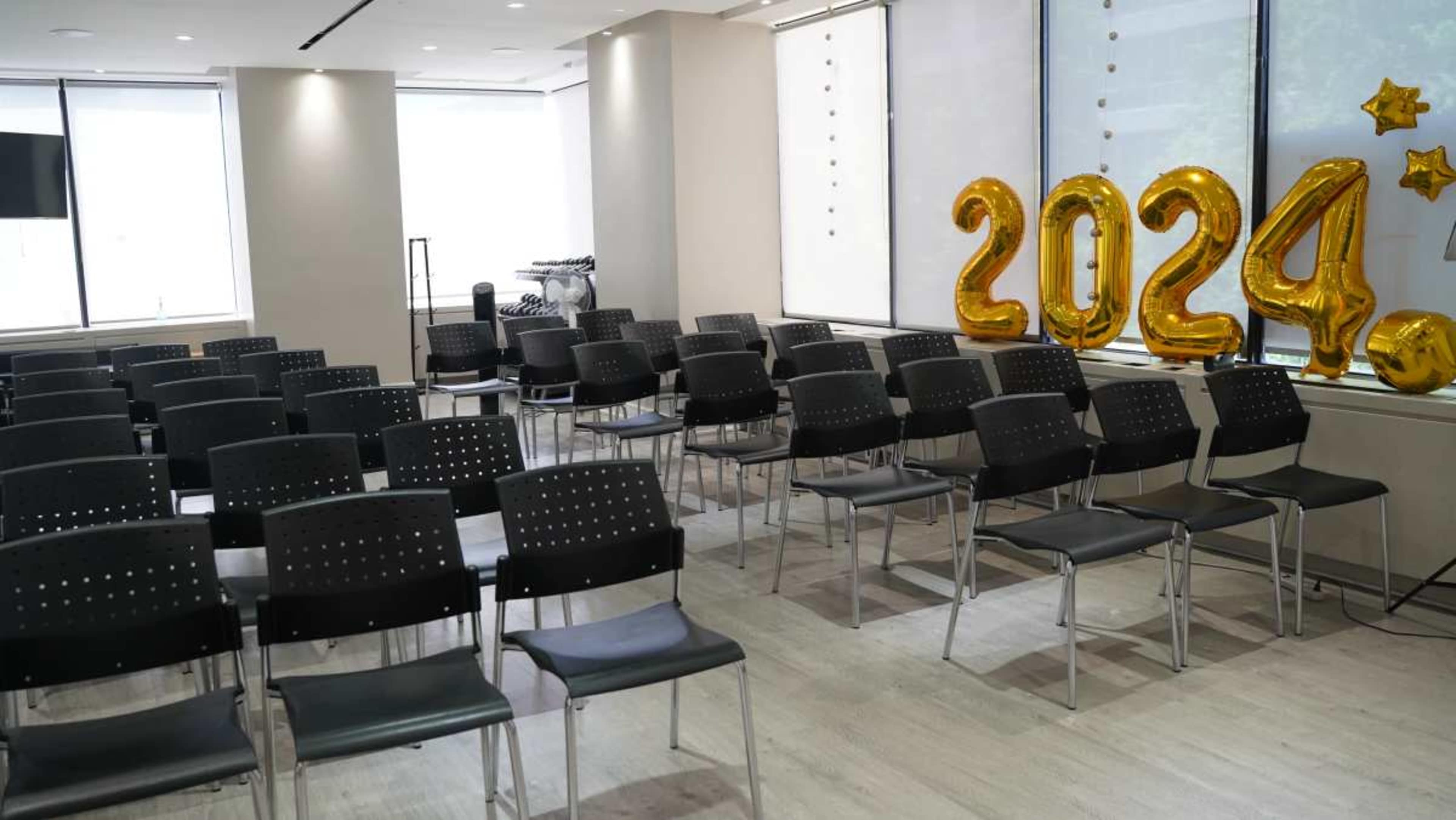 A room set up for an event features rows of black chairs and large gold balloons that read "2024."