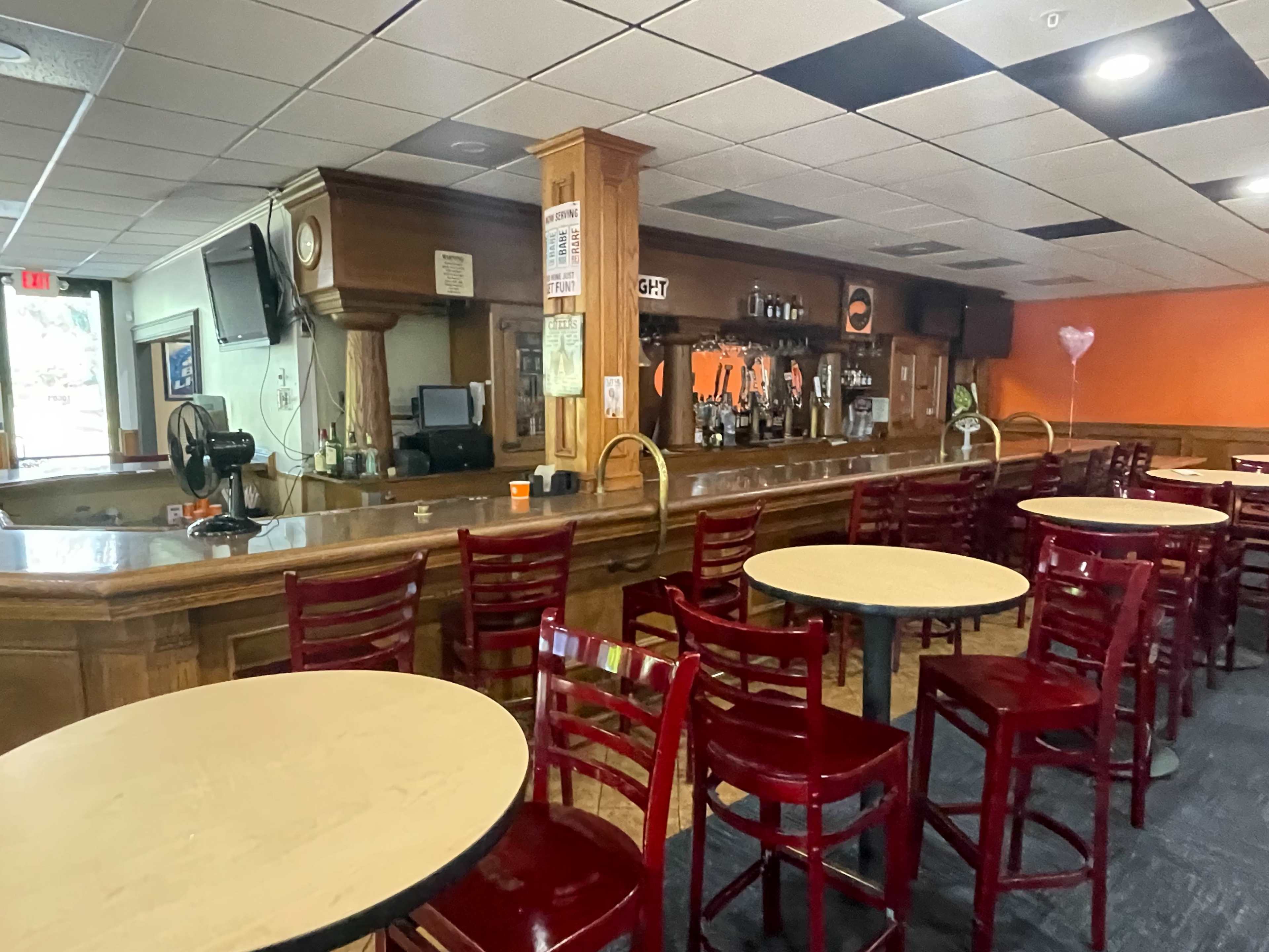 The interior of a bar features a wooden counter with empty round tables and red chairs, while shelves of bottles line the back wall.