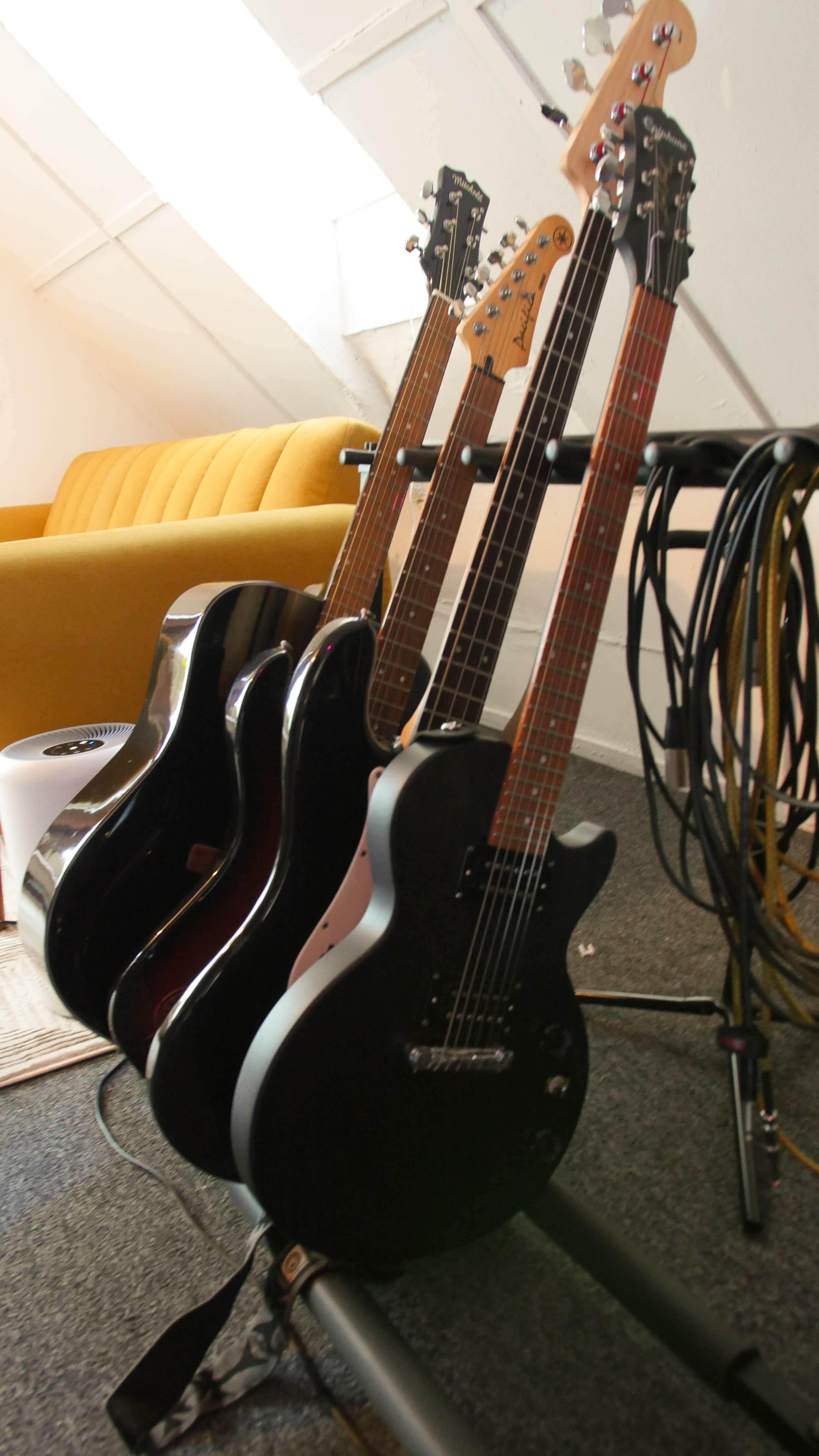 Four electric and acoustic guitars are displayed on a stand beside a yellow couch and music cables on the floor.