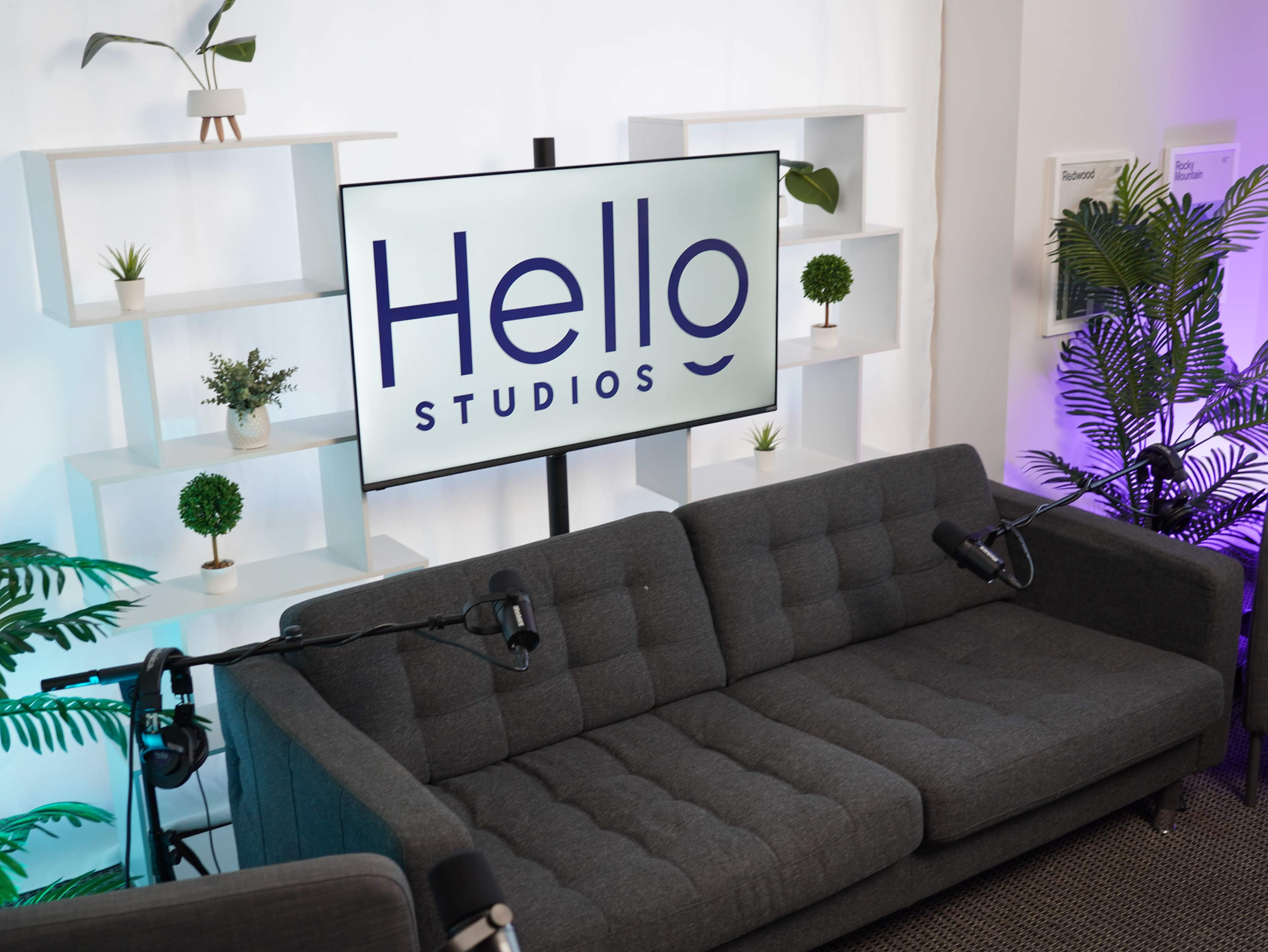 A modern studio setup featuring a gray sofa, microphones, and a large screen displaying "Hello Studios," with plants and shelves in the background.
