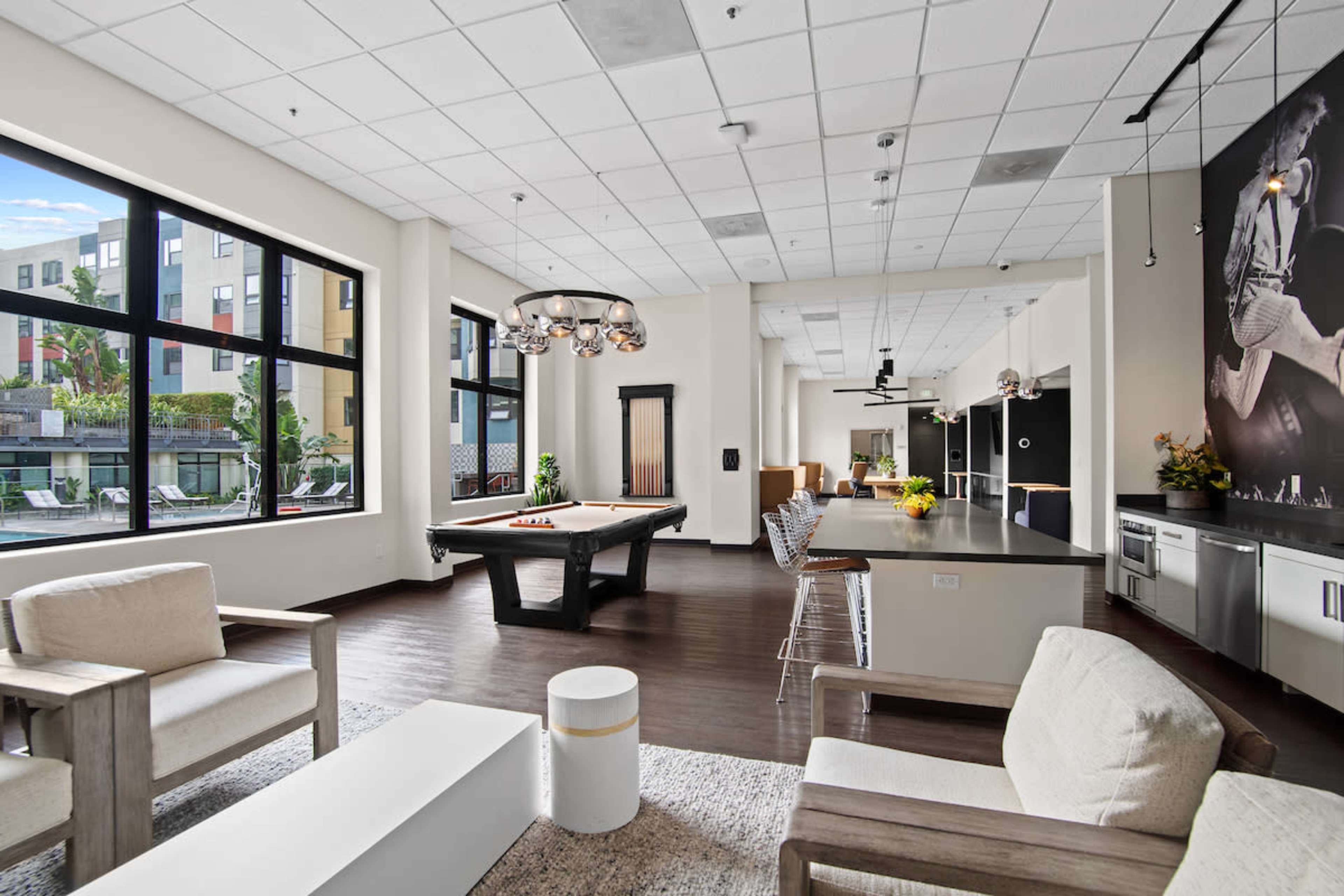 The image shows a modern lounge area featuring seating, a pool table, and large windows that provide views of an outdoor space.