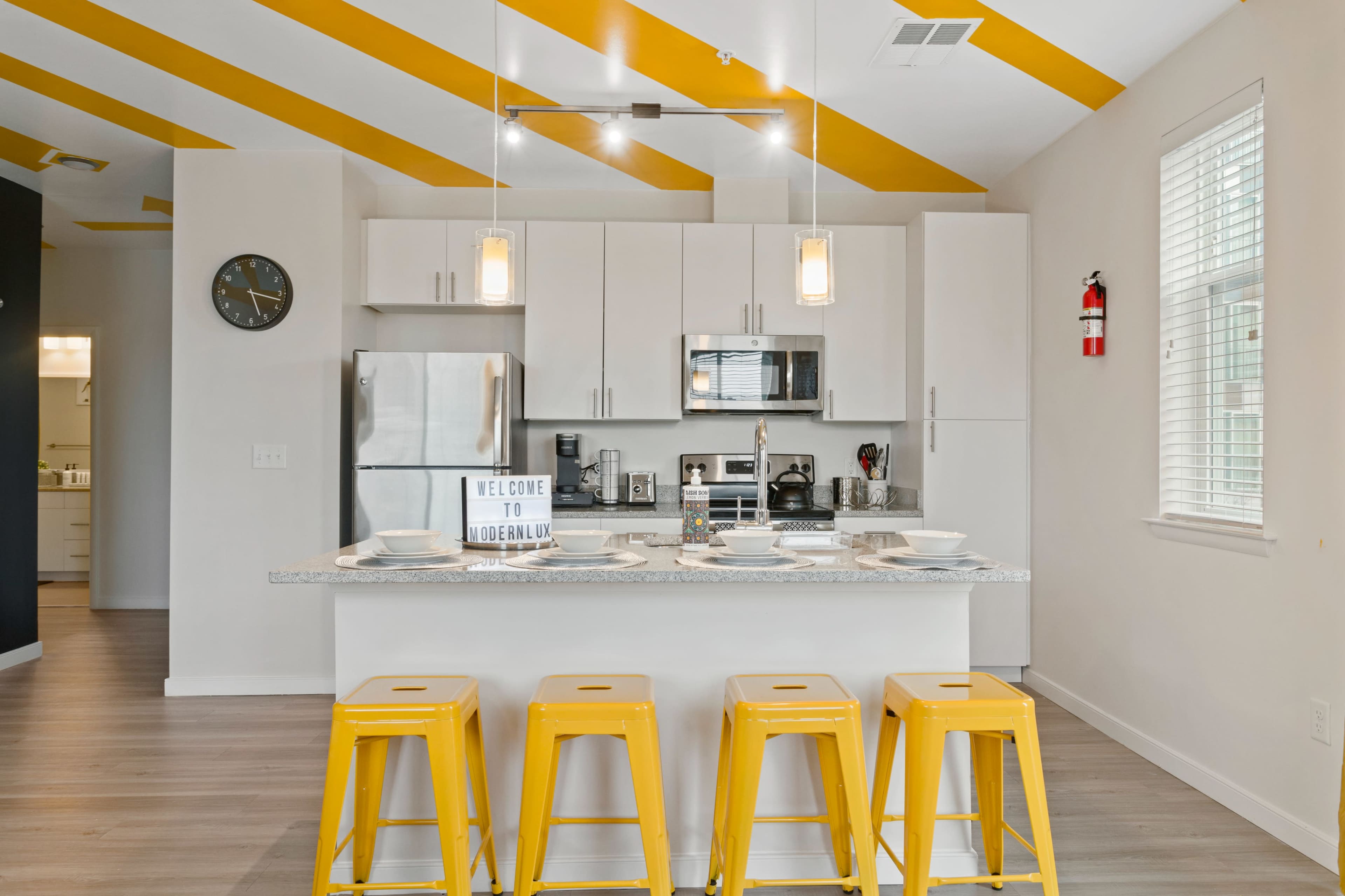 A modern kitchen features white cabinets, stainless steel appliances, and yellow bar stools at an island with a countertop.