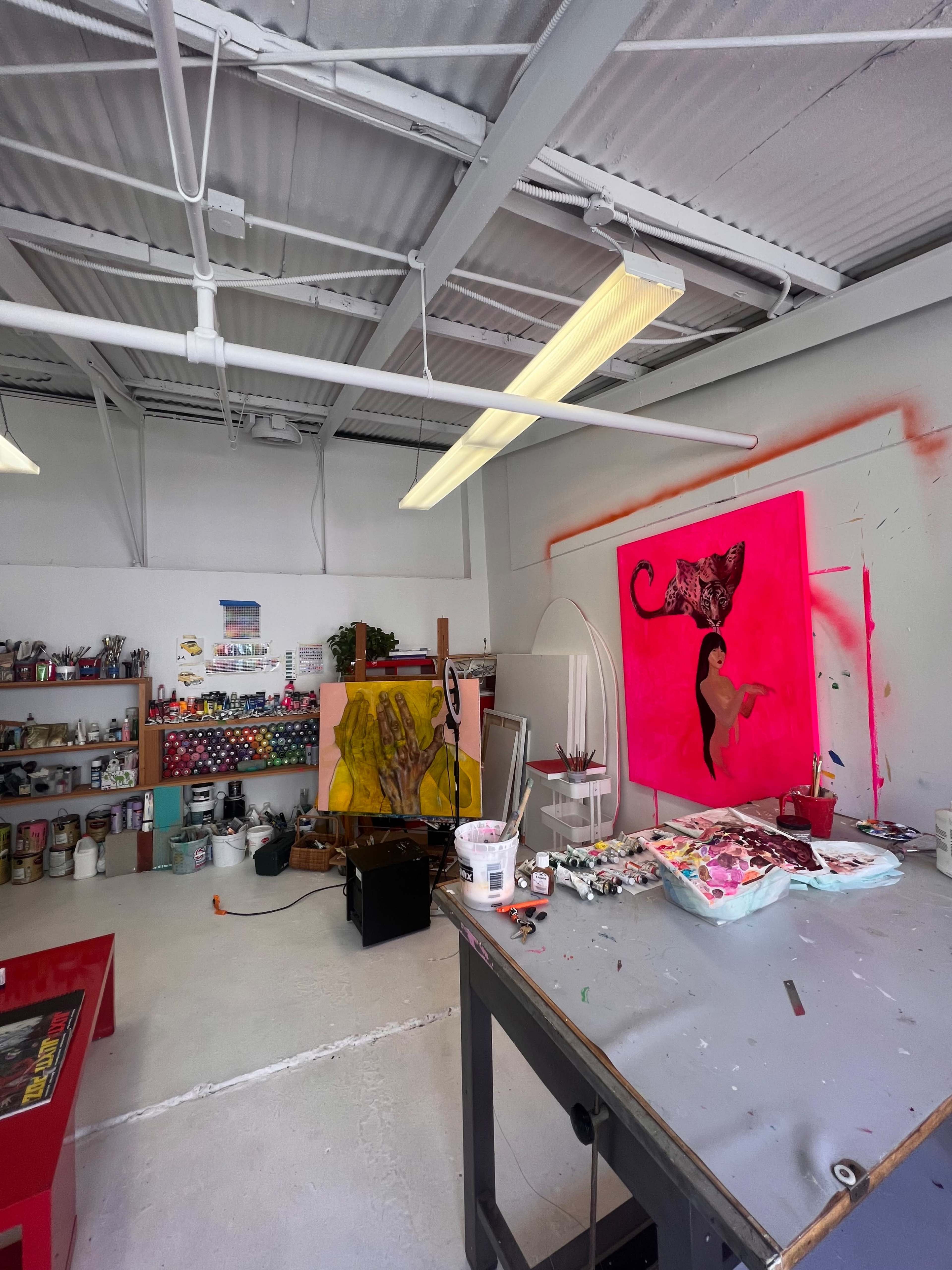 An art studio with colorful paintings, a work table scattered with supplies, and shelves filled with various art materials.