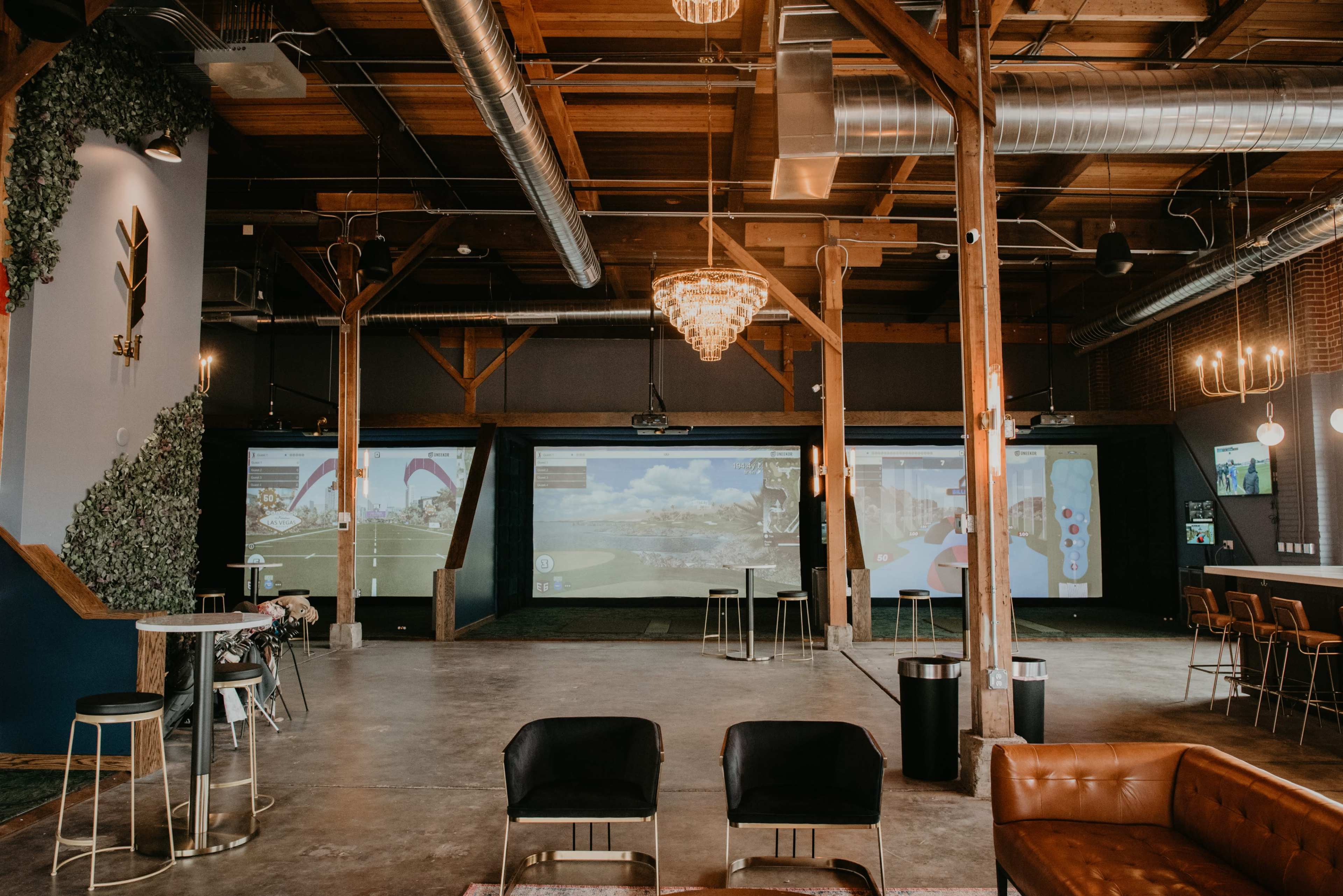 A spacious indoor venue with wooden beams, modern lighting, and multiple large screens displaying virtual golf simulations.