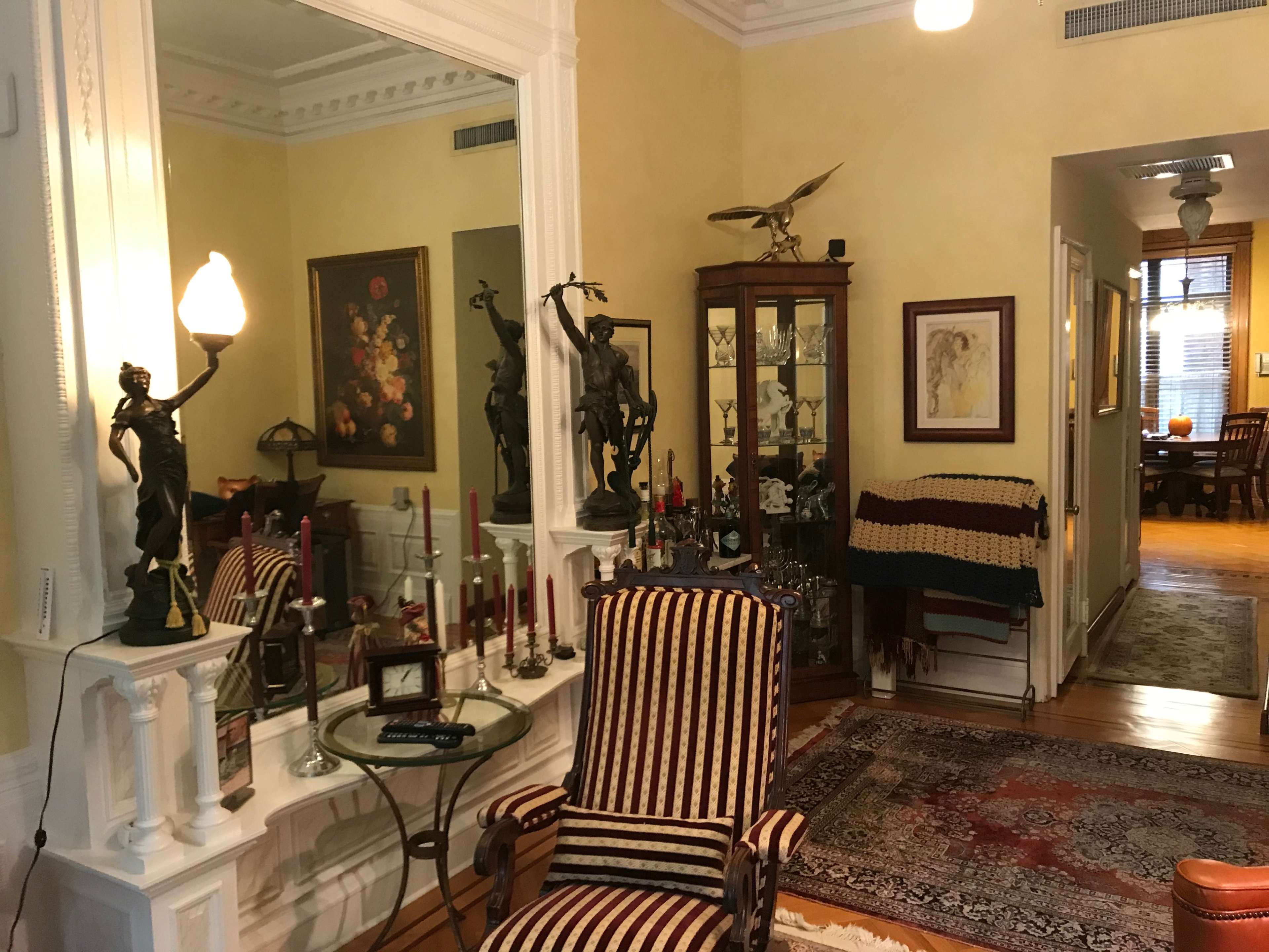 A well-furnished room with a striped armchair, a decorative mirror, a display cabinet with glass doors, and art on the walls.